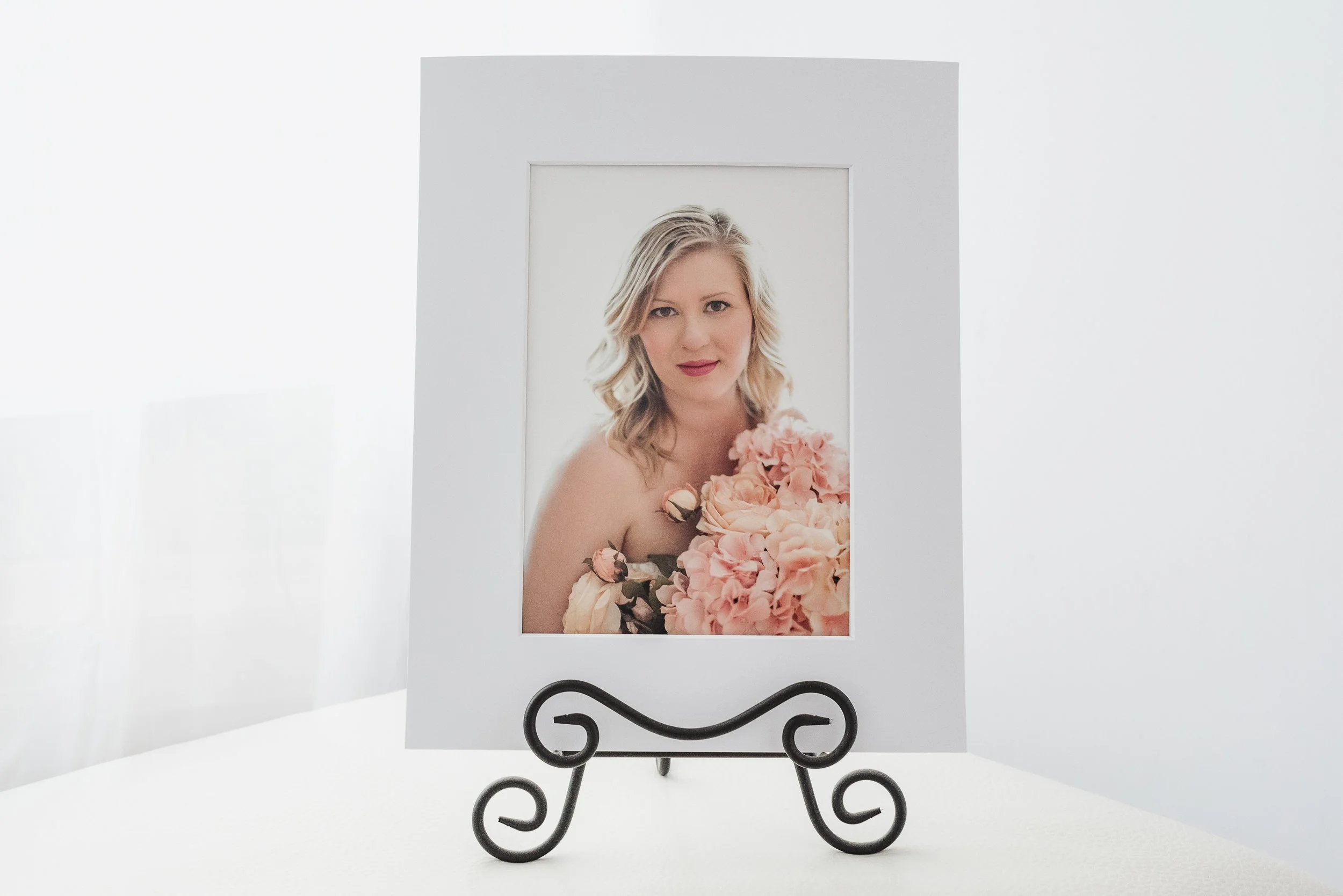 Photograph of a woman with blonde wavy hair holding a bouquet of pink and peach flowers, displayed in a white frame on a decorative black metal stand.