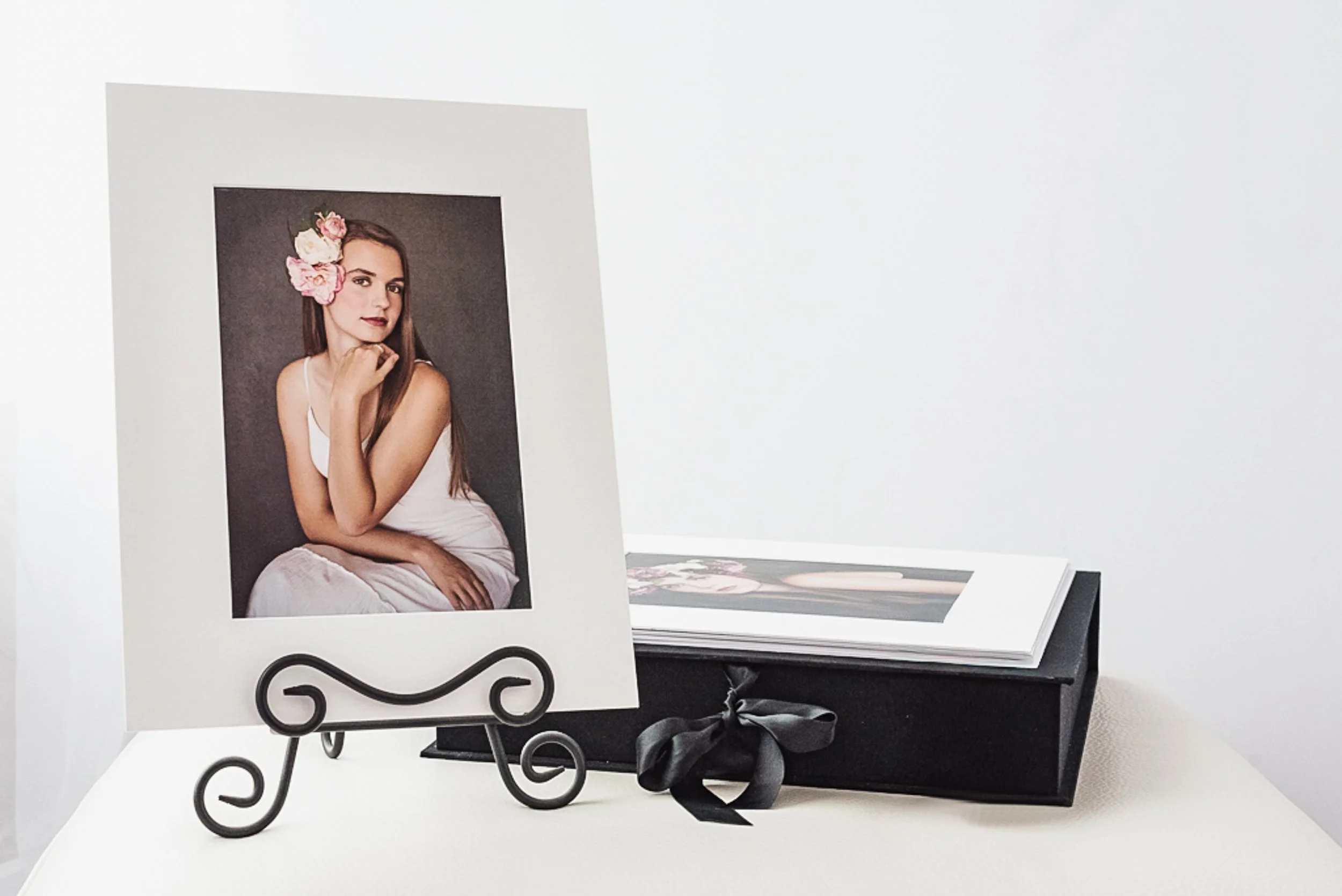 Photograph of a woman with flowers in her hair, sitting in a white dress, displayed on a small black metal stand, next to a gift box with a black ribbon, and a photo album underneath on a white surface.