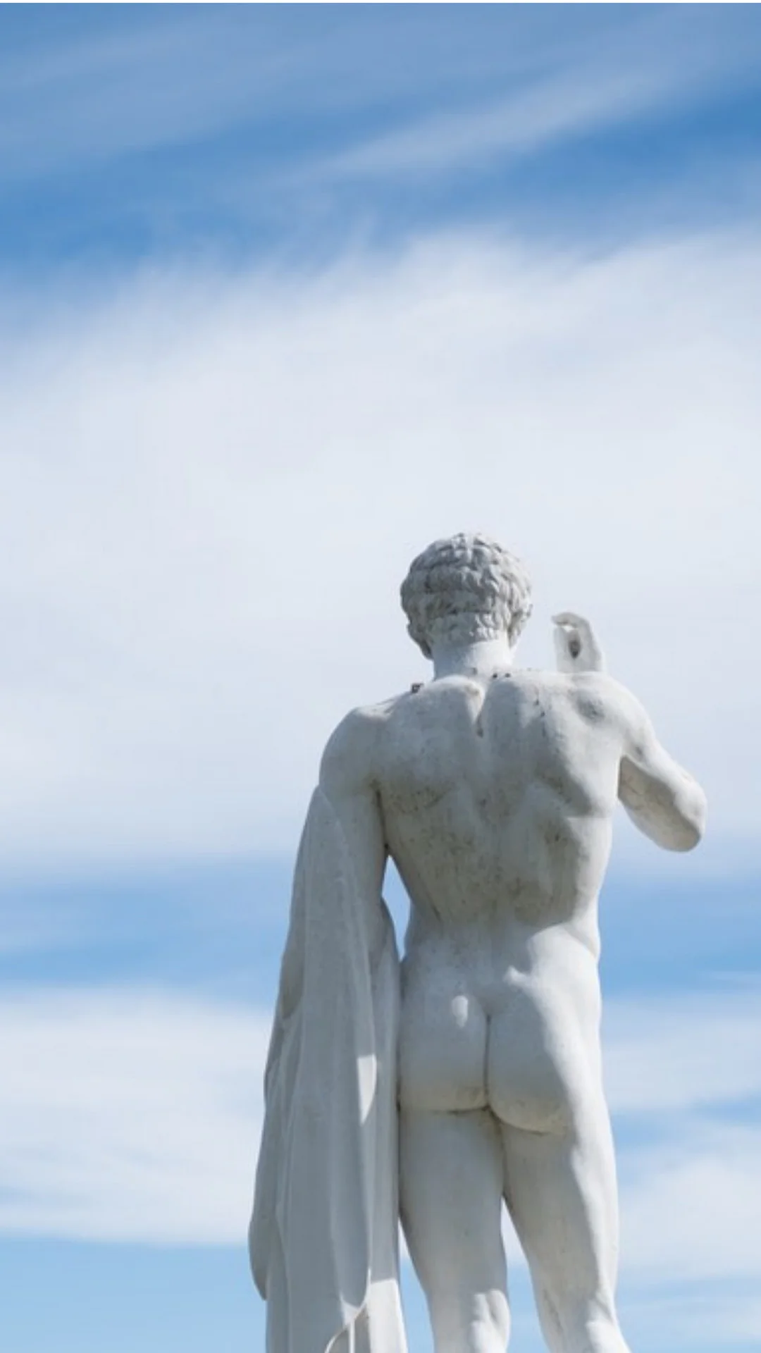 Rear view of a classical white marble statue of a muscular male athlete against a bright blue sky with wispy clouds.