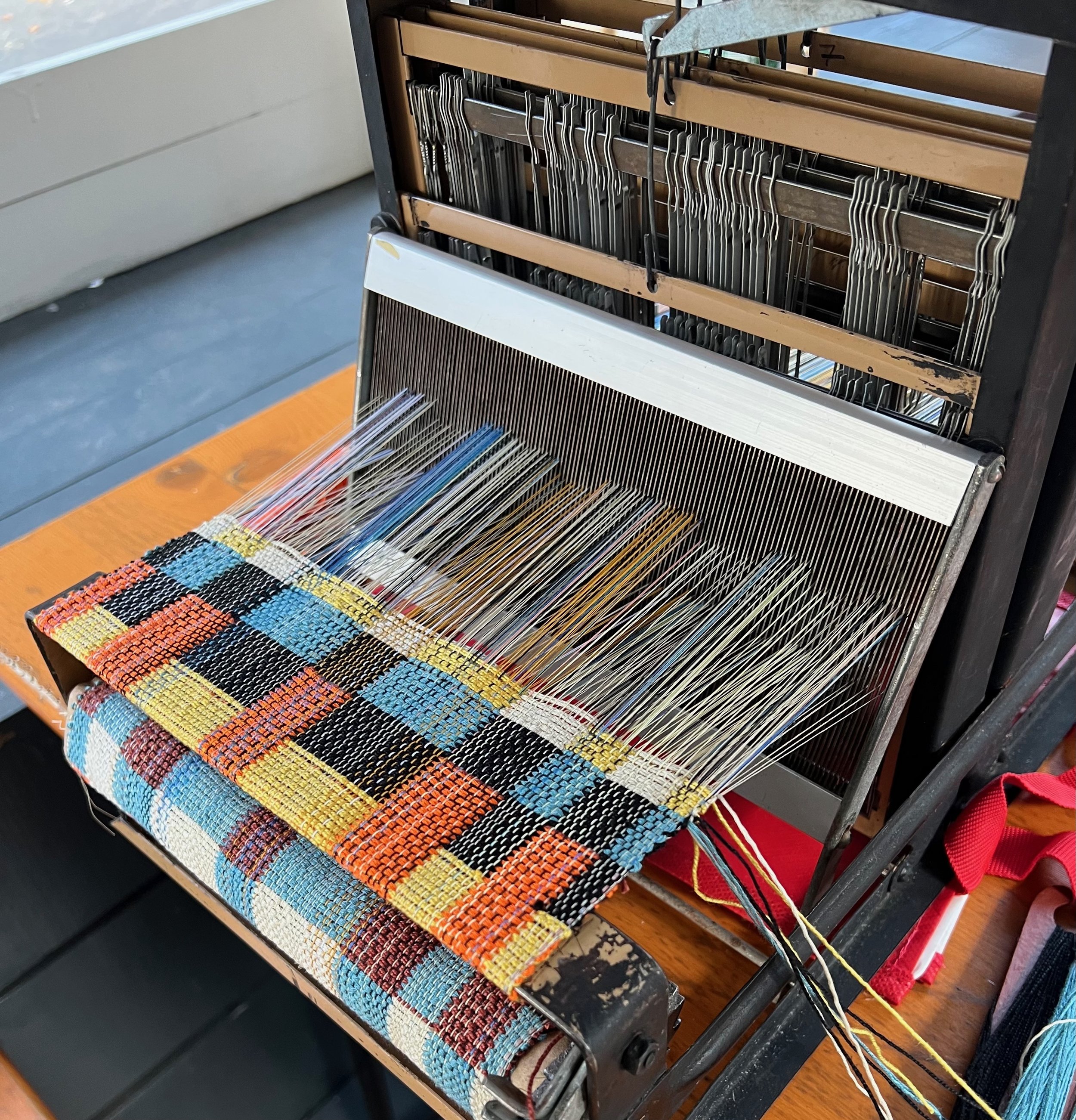 A vintage loom weaving a multicolored pattern with black, orange, yellow, blue, and white threads.