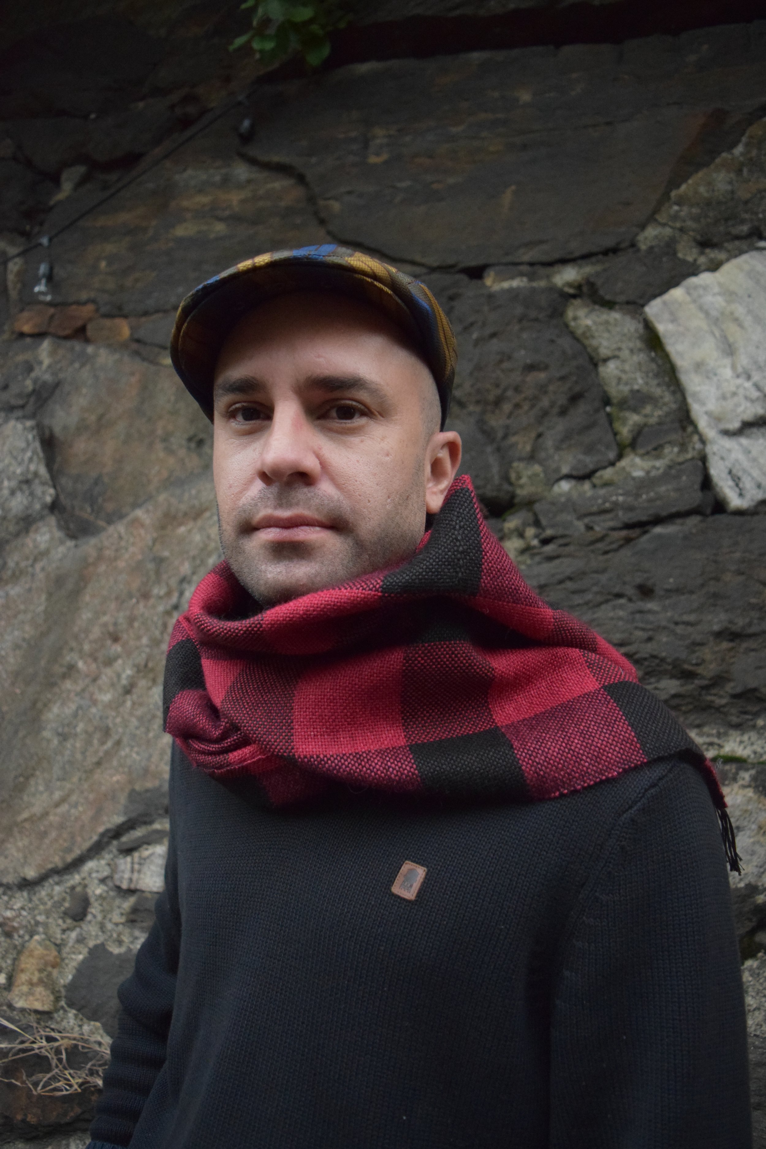 Person wearing a flat cap, red and black plaid scarf, and black sweater against a stone wall.