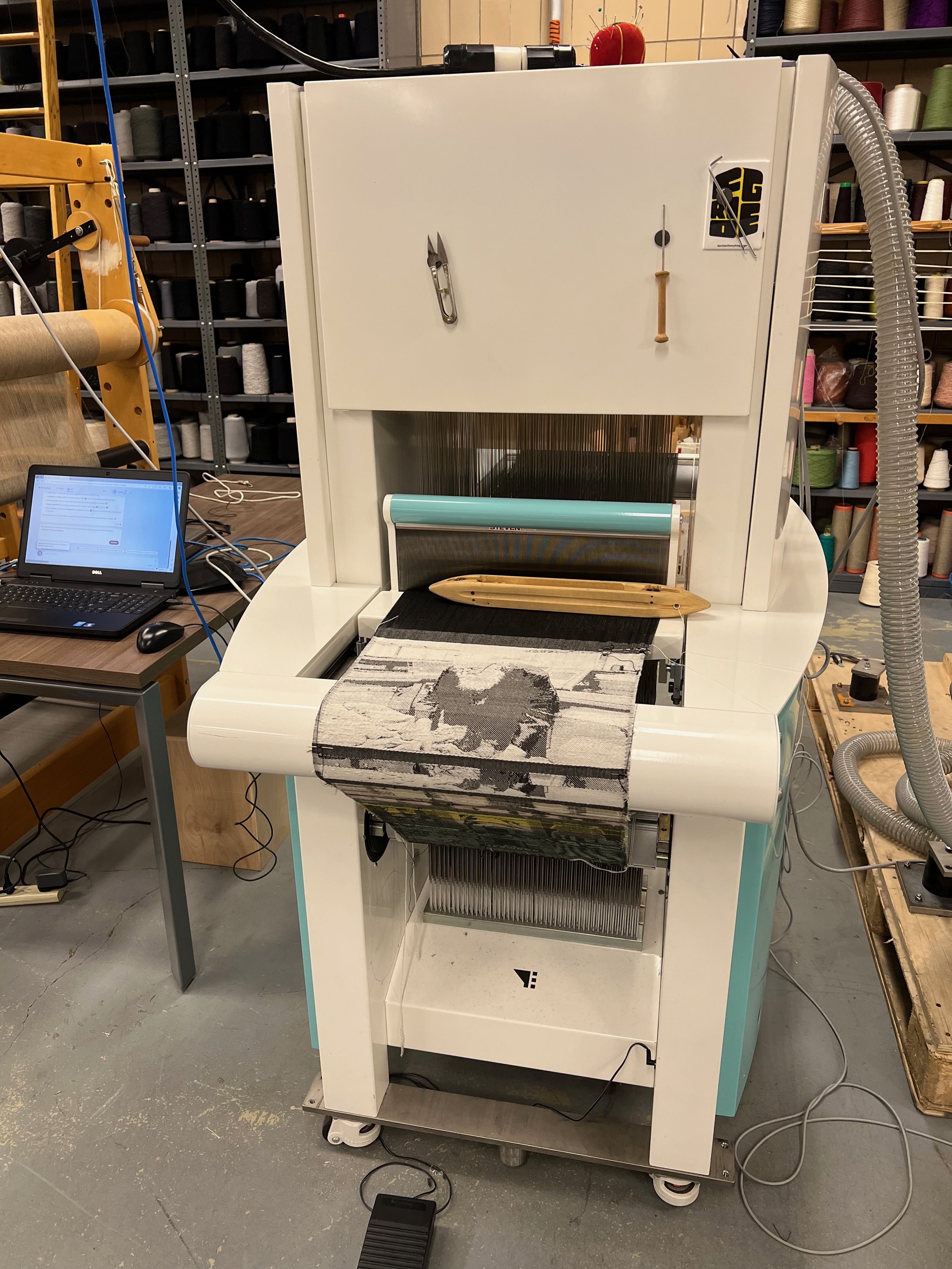 Industrial weaving machine creating patterned textile in a workshop, with spools of thread in the background and a computer nearby.