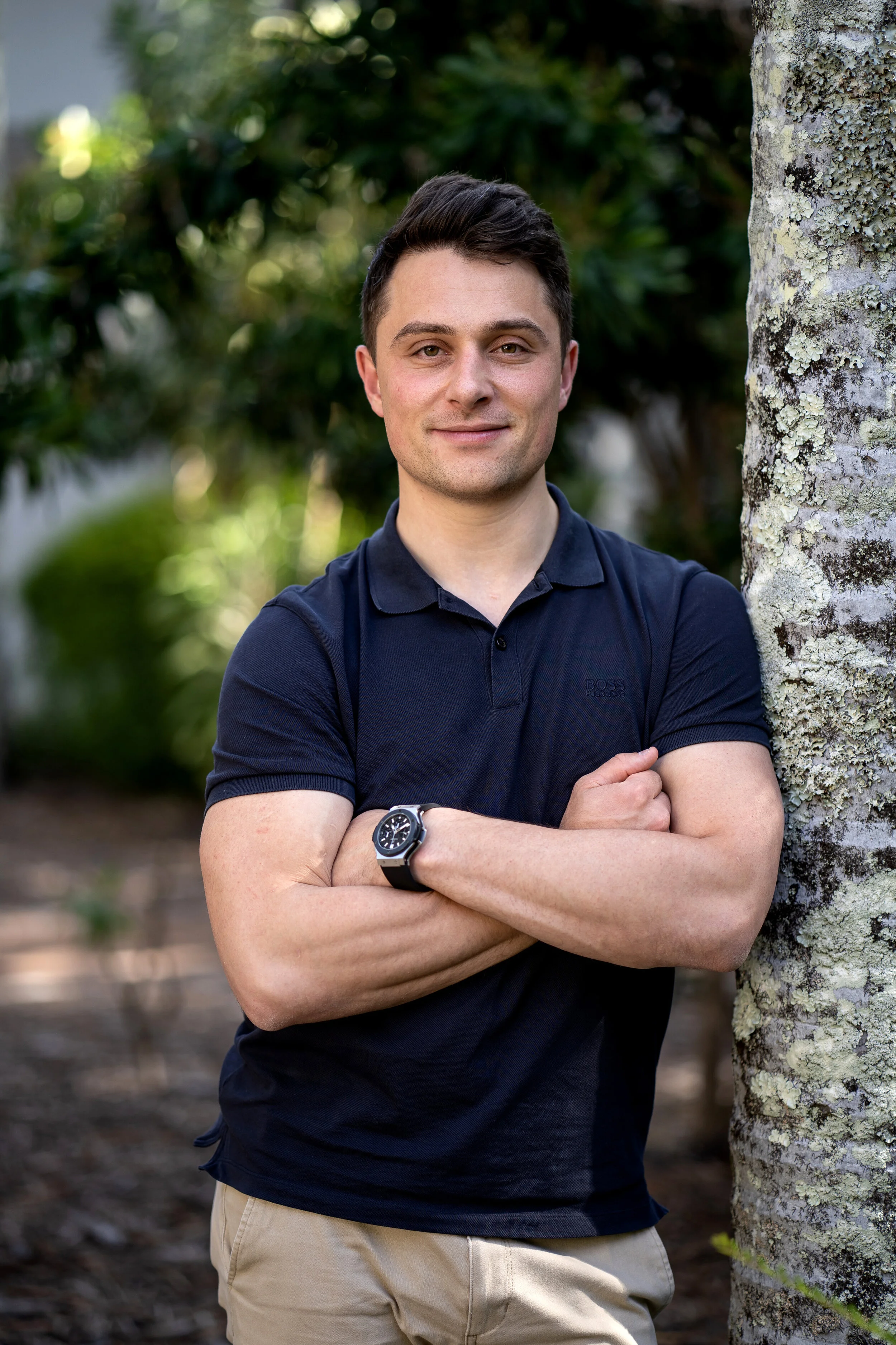 A young man with short dark hair, wearing a navy polo shirt and beige pants, standing outdoors by a tree with lichen, arms crossed, smiling at the camera with a background of green foliage.