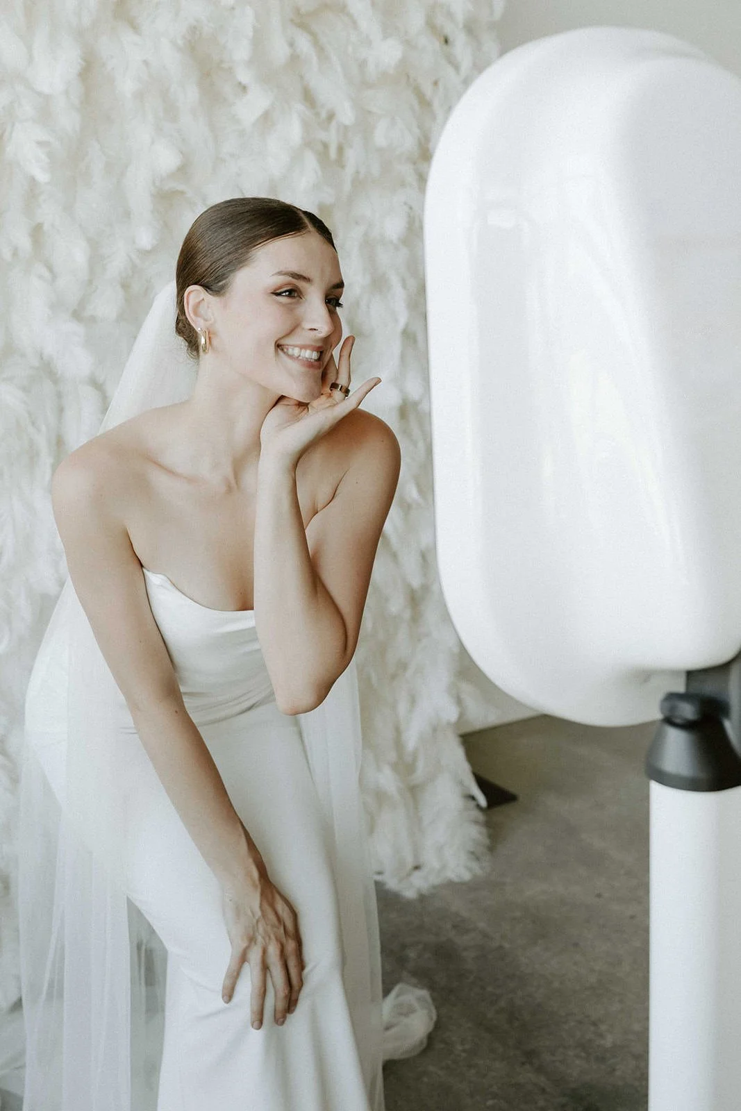 Bride smiling while using a wedding photo booth selfie mirror with a feather backdrop.