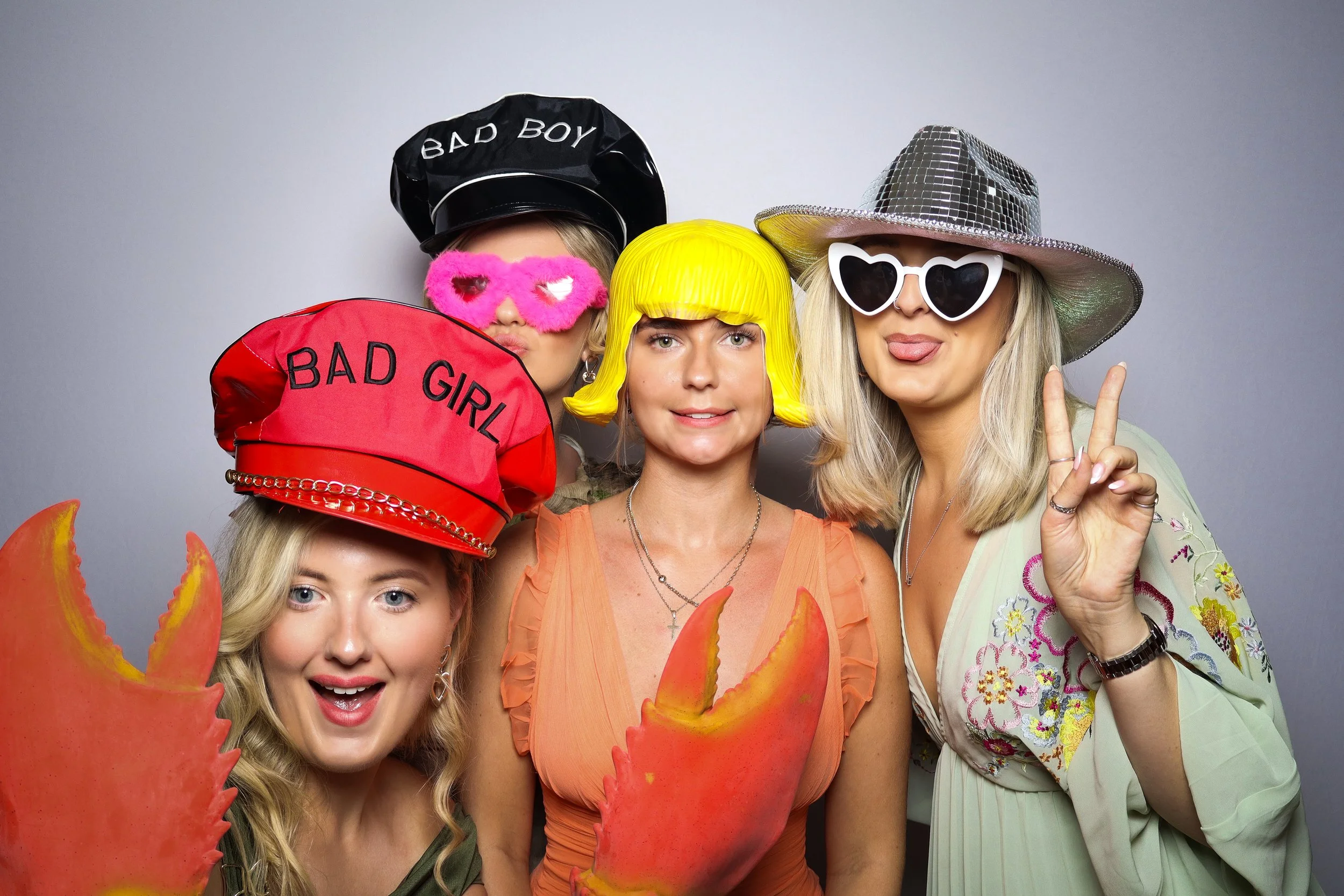 Group of four women in colorful accessories and costumes posing for a photo against a plain background. They are wearing hats, sunglasses, and holding large lobster claws as props.