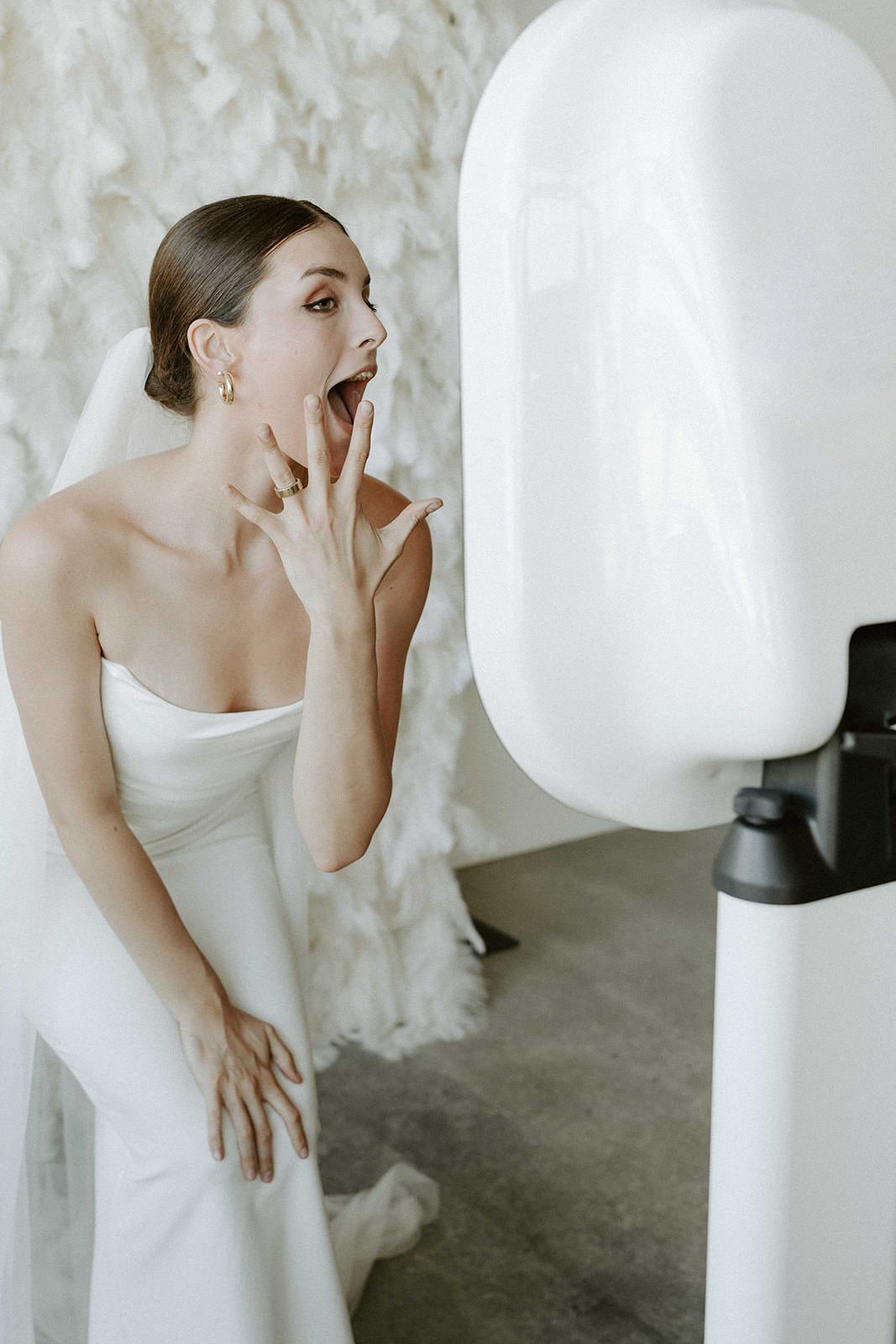A woman in a white dress, possibly a bride, looking surprised at a mirror with a feathered backdrop.