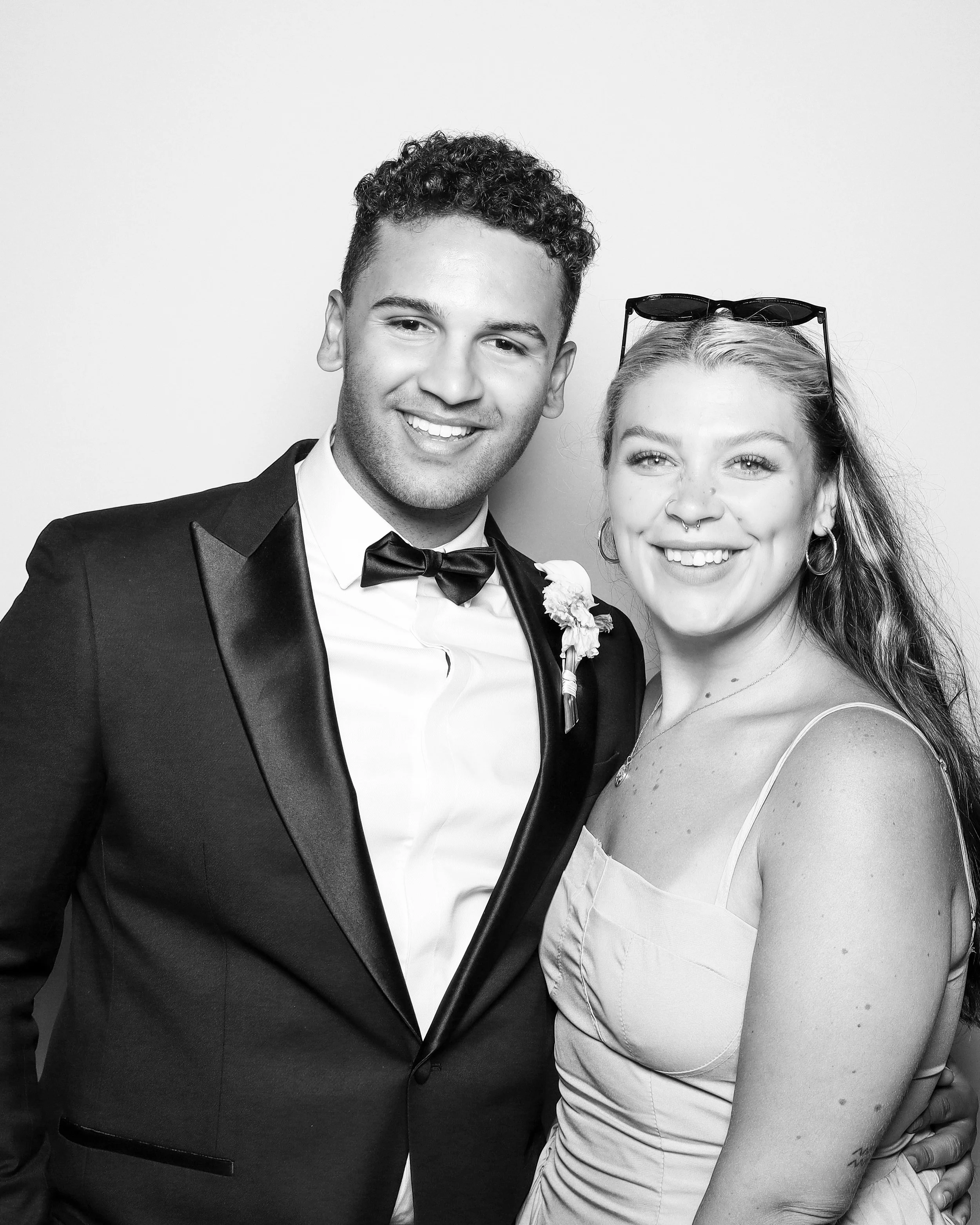 A black and white photo of a man and a woman close together, smiling at the camera. The man is dressed in a tuxedo with a bow tie, and the woman has sunglasses on her head, a septum pierced nose, and earrings, wearing a silky dress.