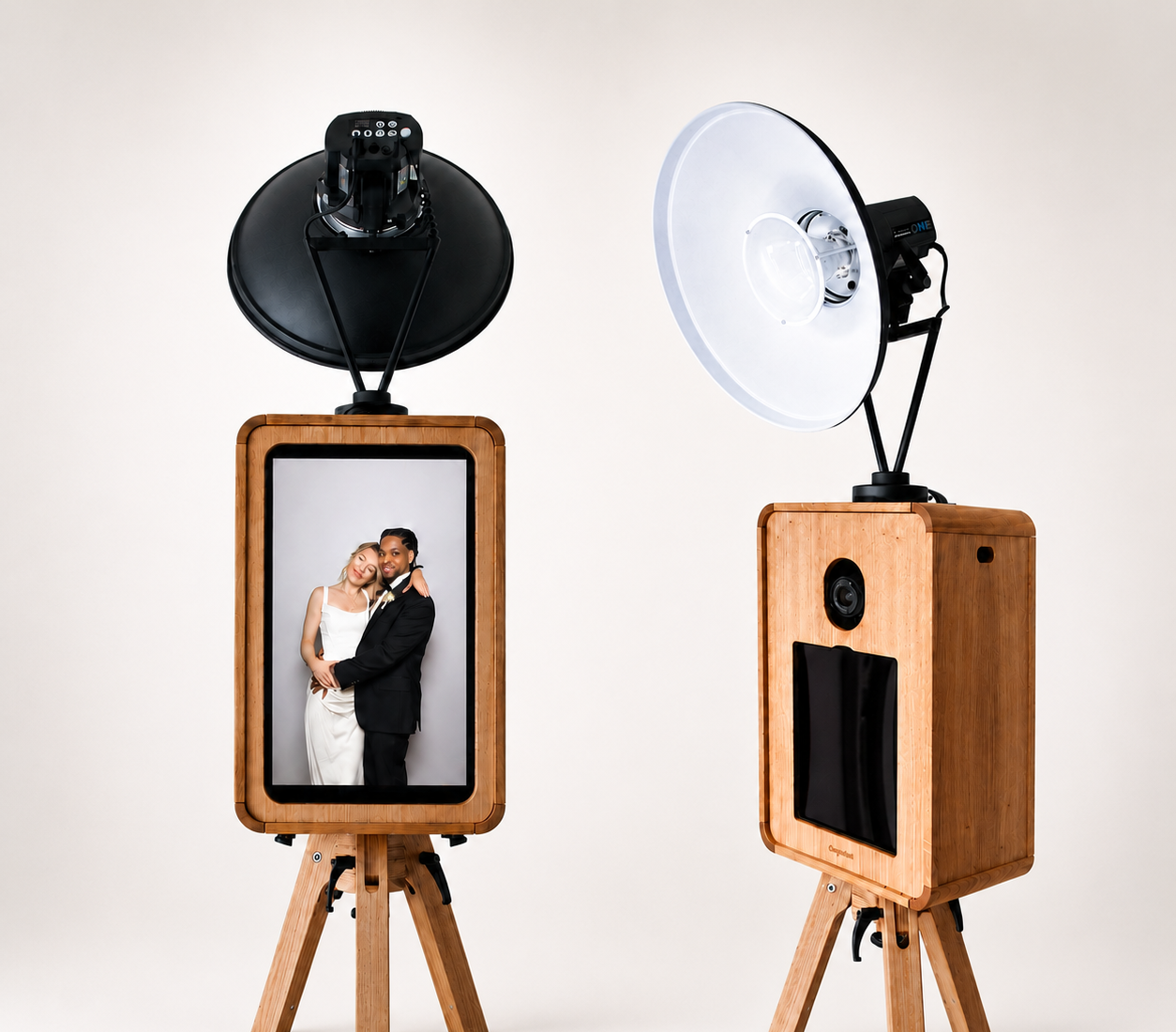 Two wooden photo booths with large studio lights above, one displaying a photo of a smiling woman in a white dress and a man in a black tuxedo, in front of a light gray background.