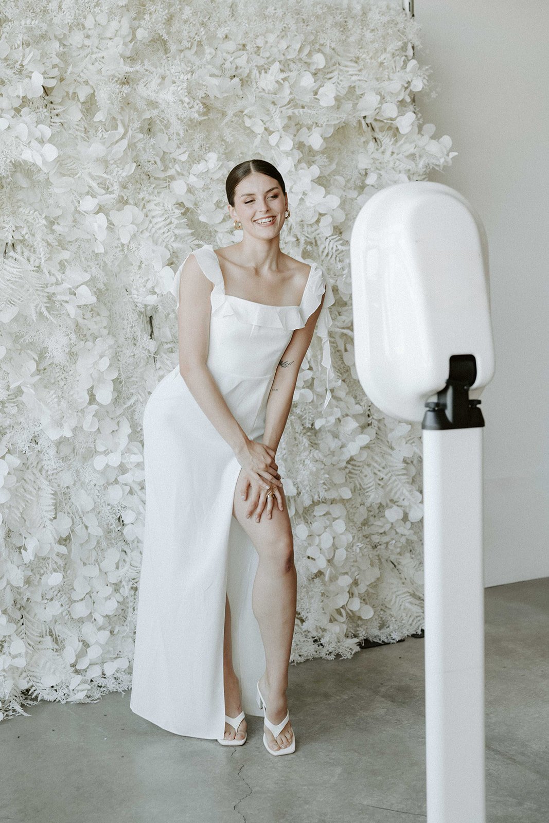 Woman in white dress posing in front of floral backdrop with a photo booth.