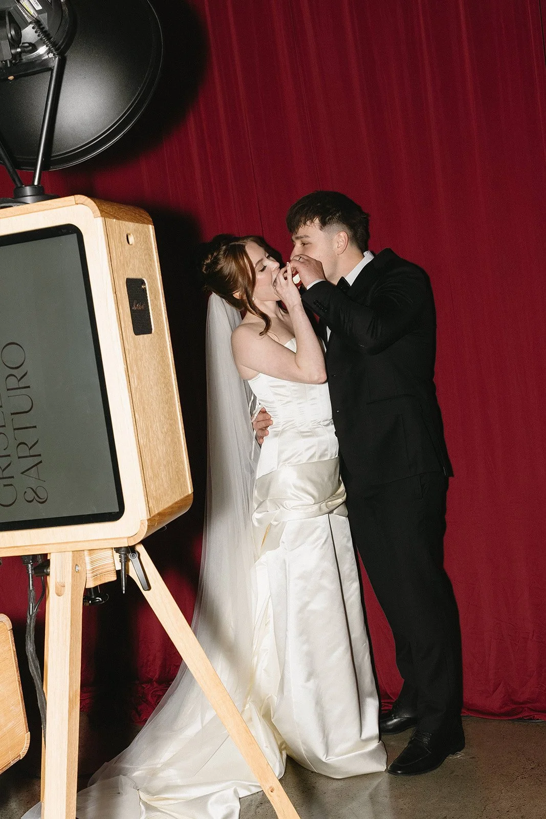 A bride and groom sharing a romantic moment during their wedding photoshoot in front of a deep red curtain, with a photo booth visible in the foreground.