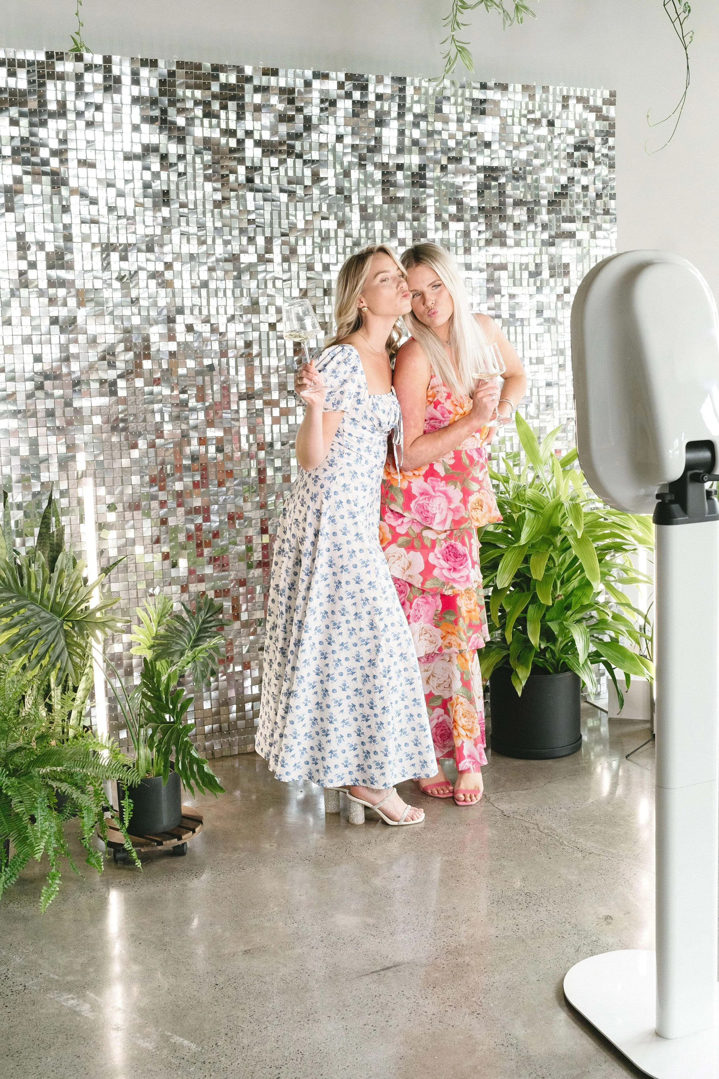 Two women in floral dresses posing and making kissy faces while holding glasses of wine in front of a metallic mosaic wall, surrounded by green plants
