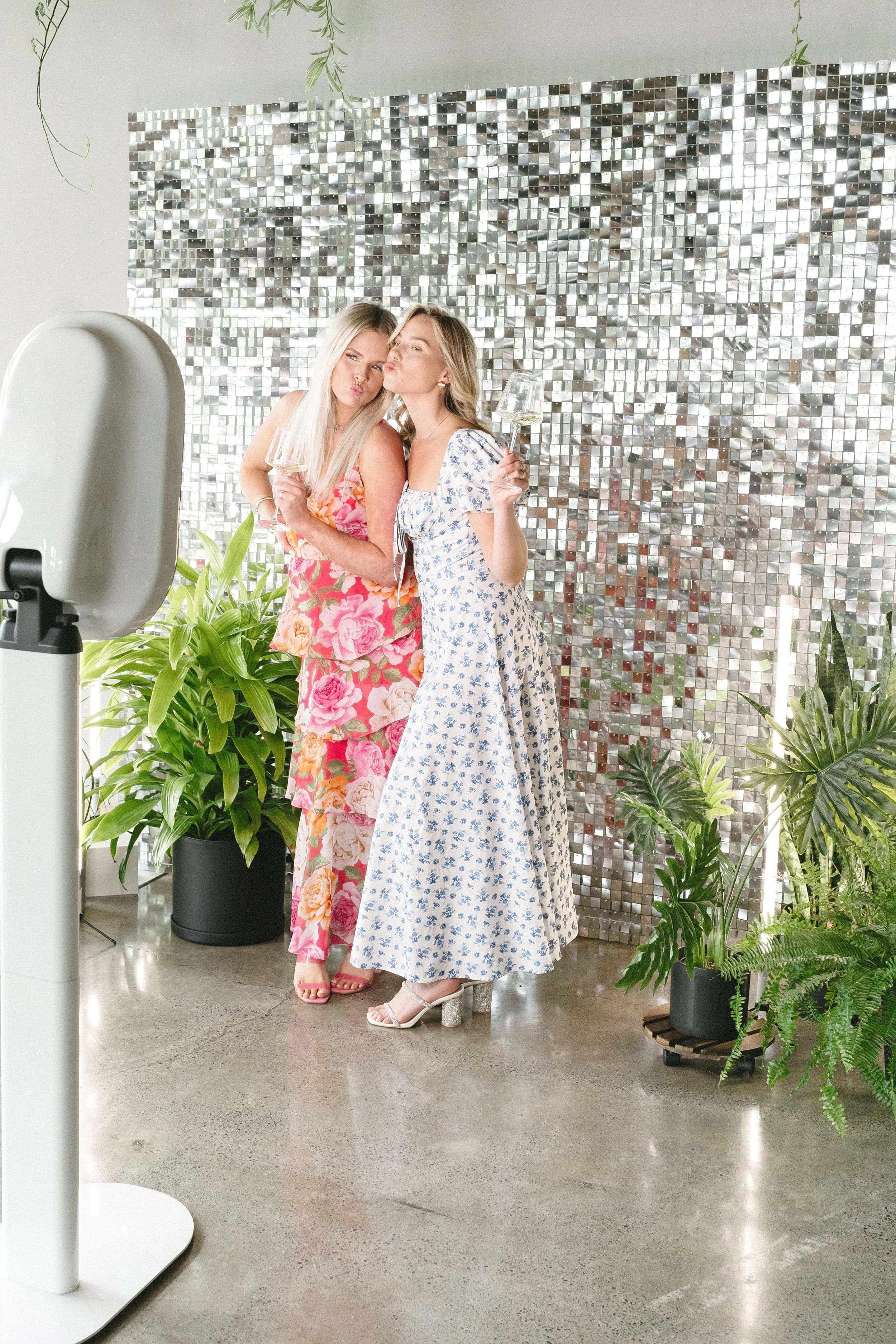 Two women in floral dresses posing with wine glasses in front of a silver sequin backdrop, surrounded by plants, using a photo booth.