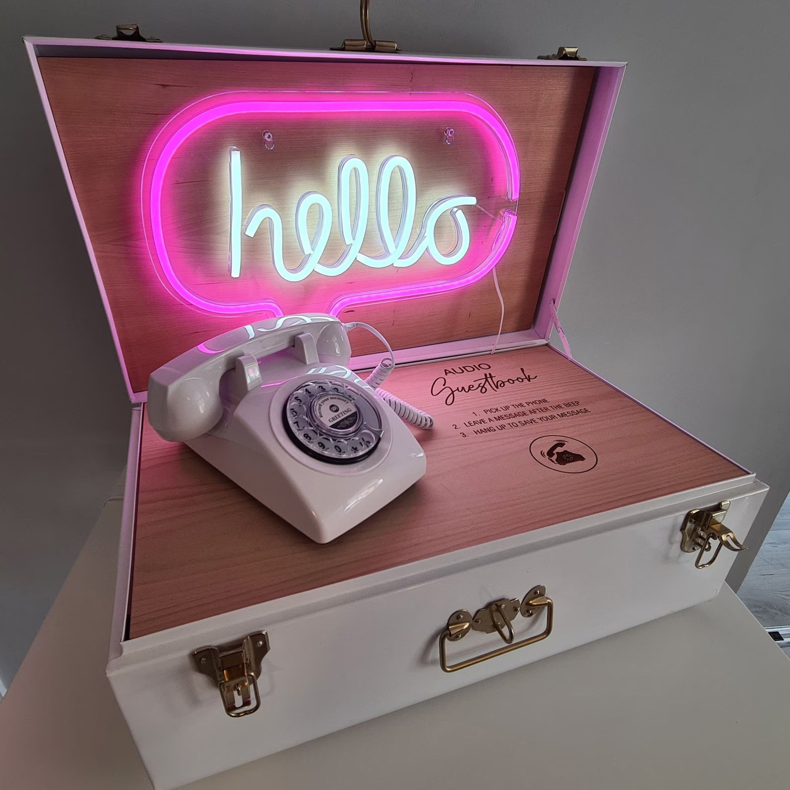 A white vintage rotary phone placed inside a decorated wooden box with a pink neon sign that says 'hello'. The box has pink interior and instructions for an audio guestbook feature.