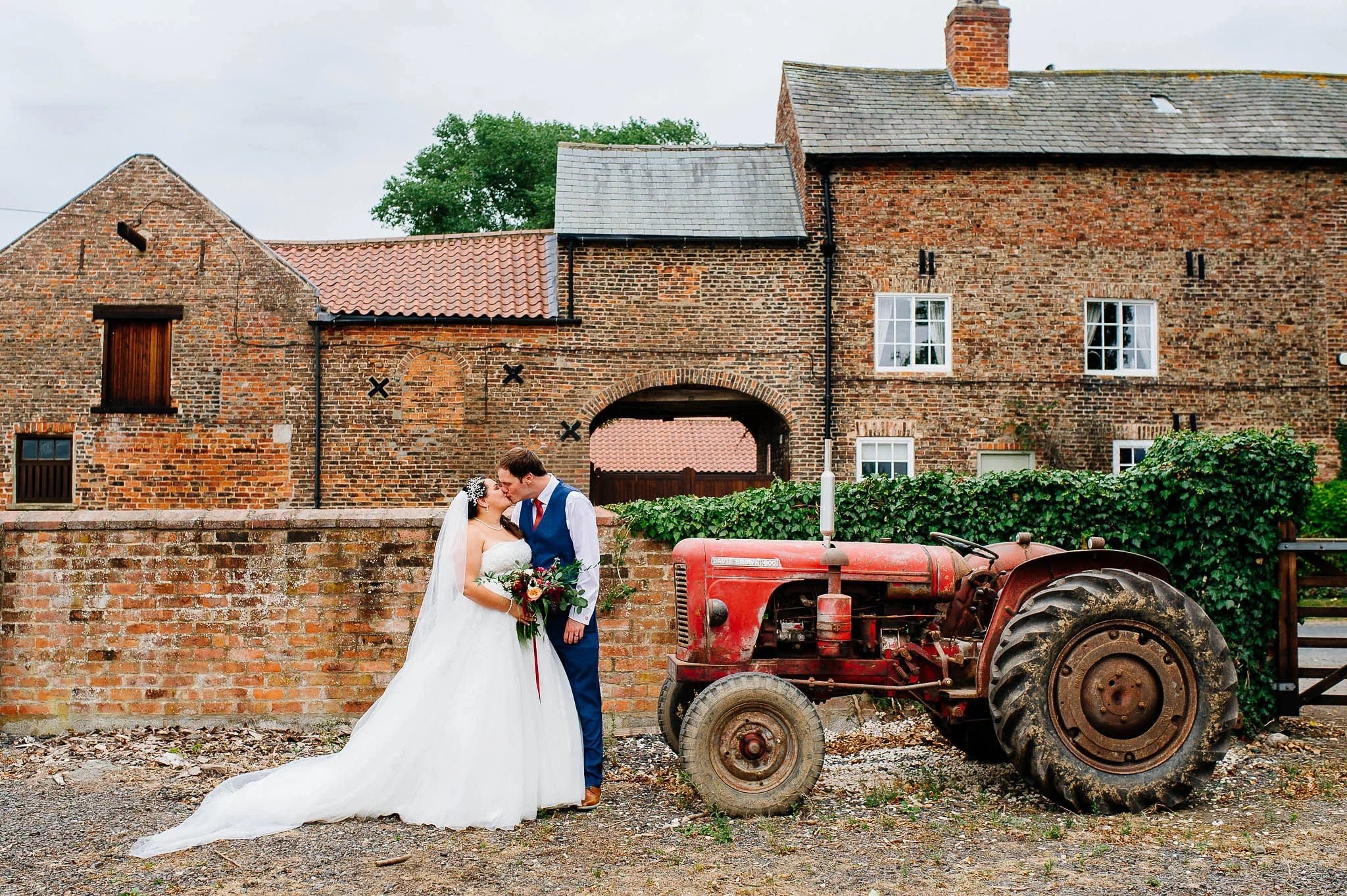 Bride and groom standing by a vintage red tractor outside The Normans wedding venue in York.