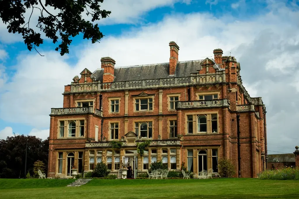 Exterior of Rossington Hall with red-brick facade, large windows, manicured gardens and blue sky.