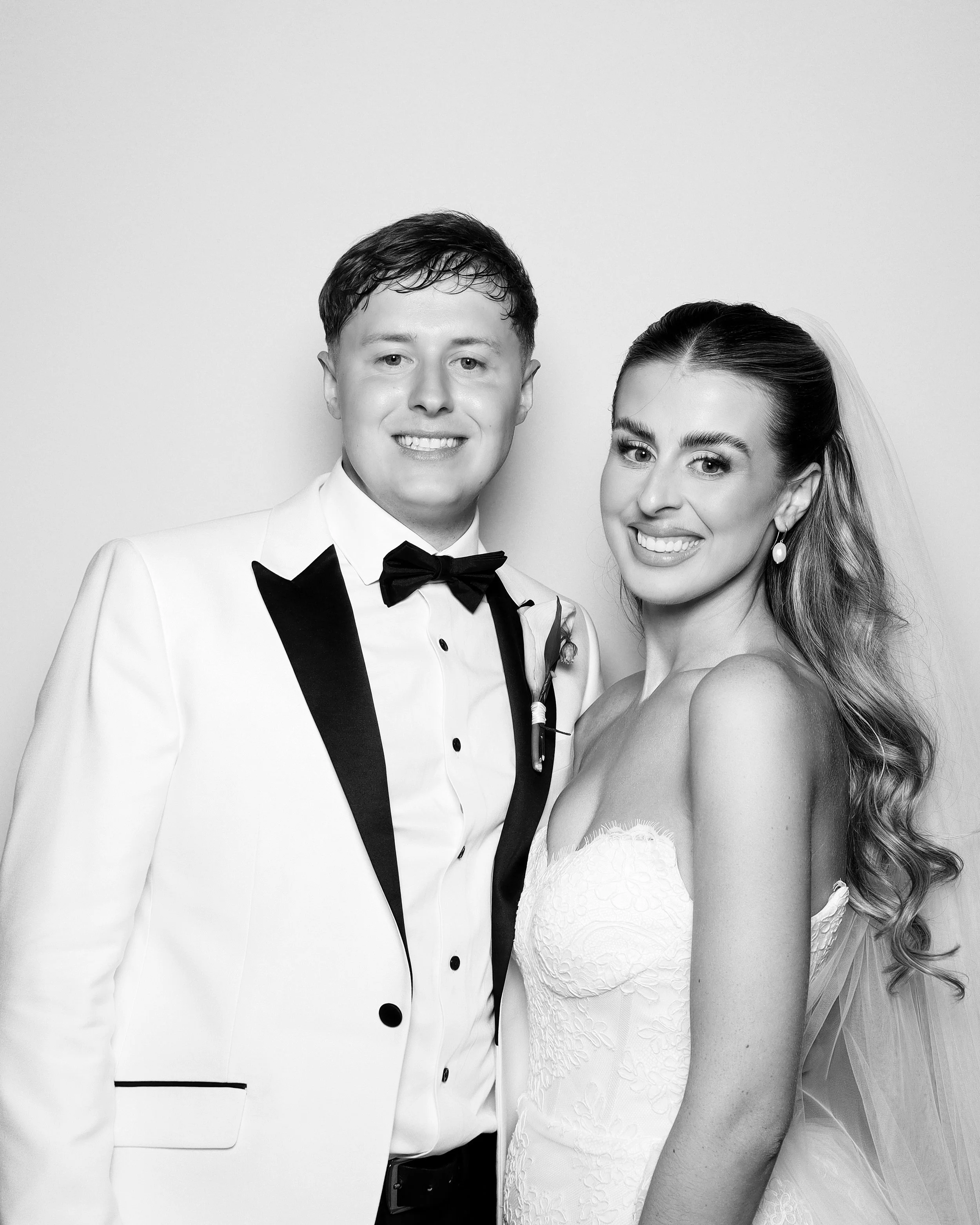 A black and white photo of a couple, dressed in formal attire, smiling and looking at the camera. The man is wearing a tuxedo with a bow tie, and the woman is in a strapless dress with earrings, with her hair styled and a veil visible.