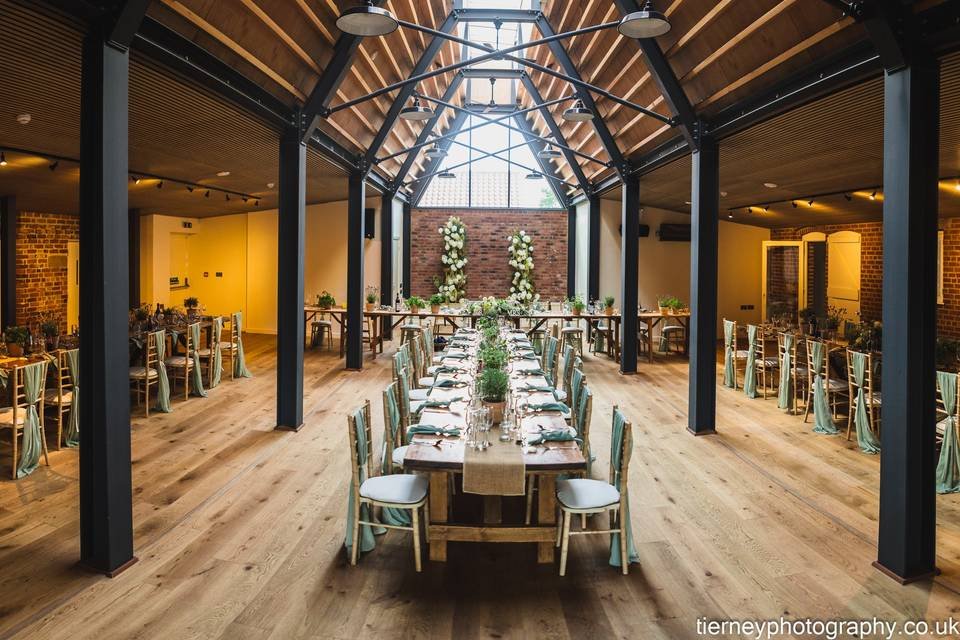 Elegant event space with long wooden tables set for dining, decorated with green plants, in a bright room with brick and wood accents.