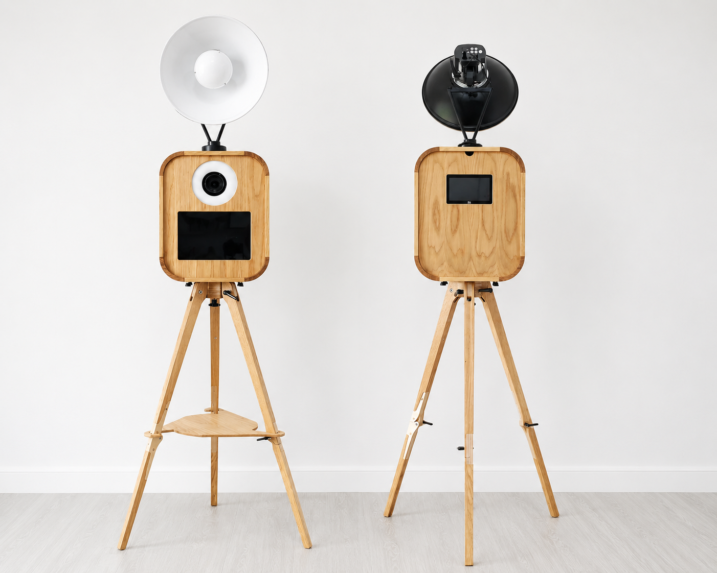 Two wooden camera-style structures on tripod stands with screens and camera lenses, one with a white dome light and the other with a black umbrella light, against a plain white wall.