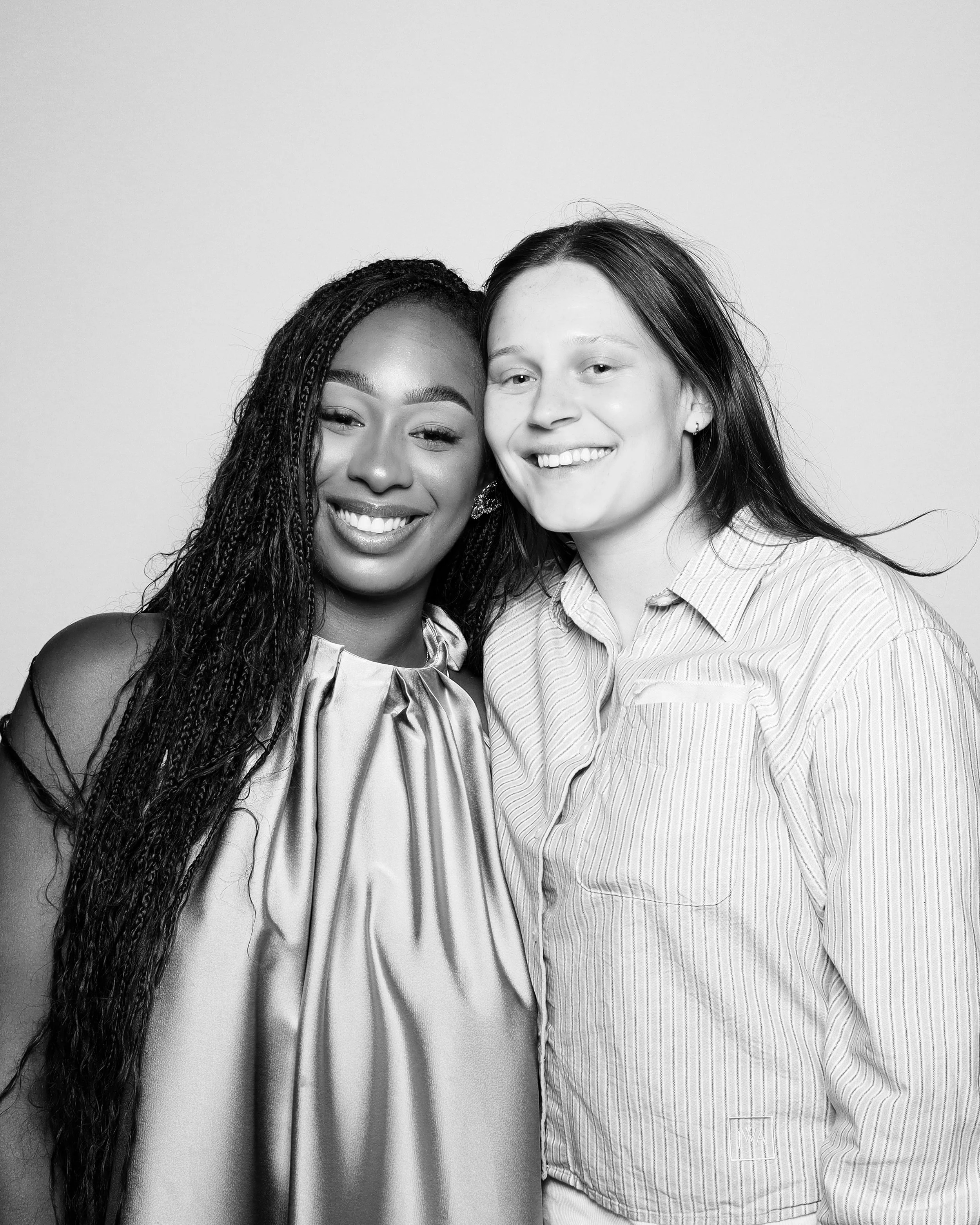 Two women smiling with heads close together, one with long braided hair and the other with straight dark hair, both wearing striped clothing, in a studio setting.