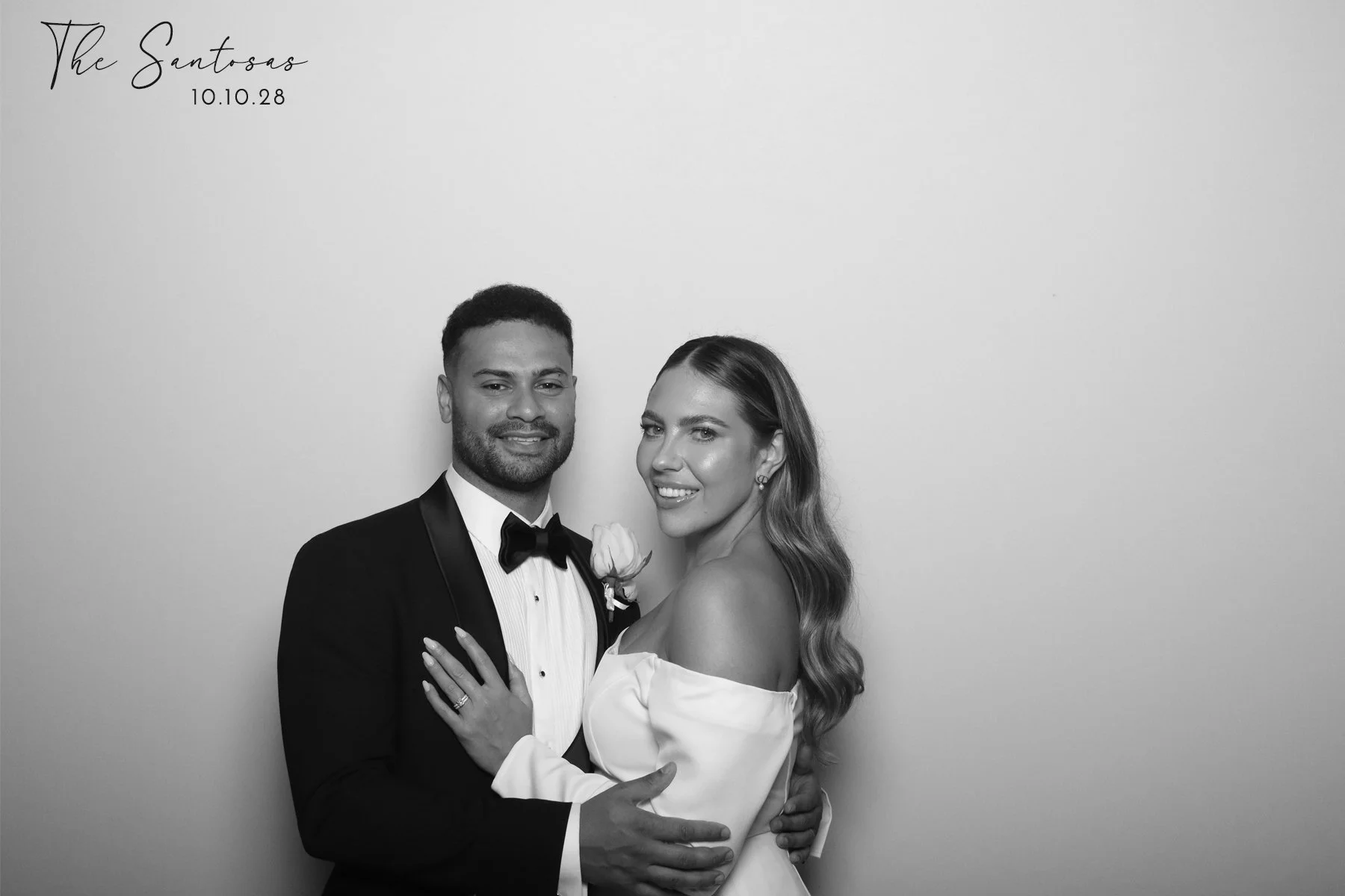Black and white photo of a smiling couple dressed in formal wedding attire, with the man wearing a tuxedo and the woman in a wedding dress, standing close together against a plain background. Text in the upper left corner reads 'The Santosas 10.10.28