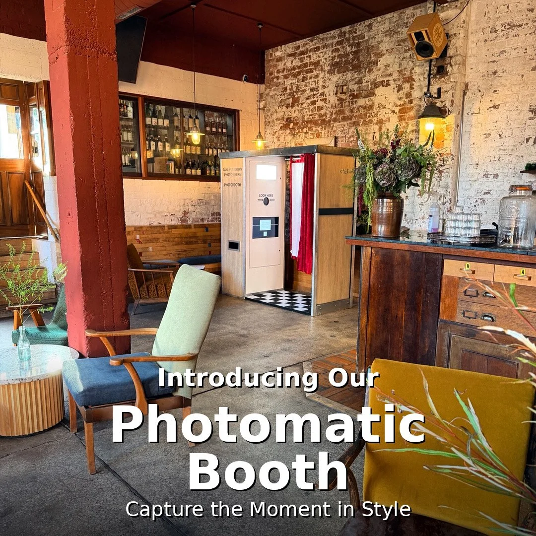 Interior of a cozy cafe with vintage furniture, plants, a photo booth, and a brick wall. The photo booth is located in the center against the wall and has a red curtain door. The scene includes a brown cabinet with glass jars and a large flower arrangement.