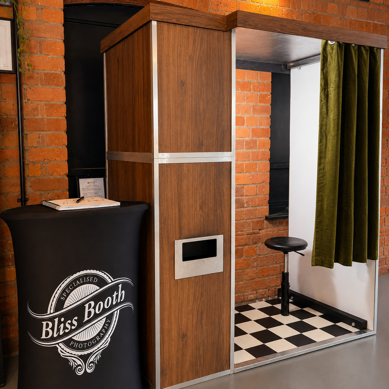 Photo booth area with a black curtain, a stool, a green curtain, and a wood-paneled enclosure labeled 'Photomatic' against a brick wall.
