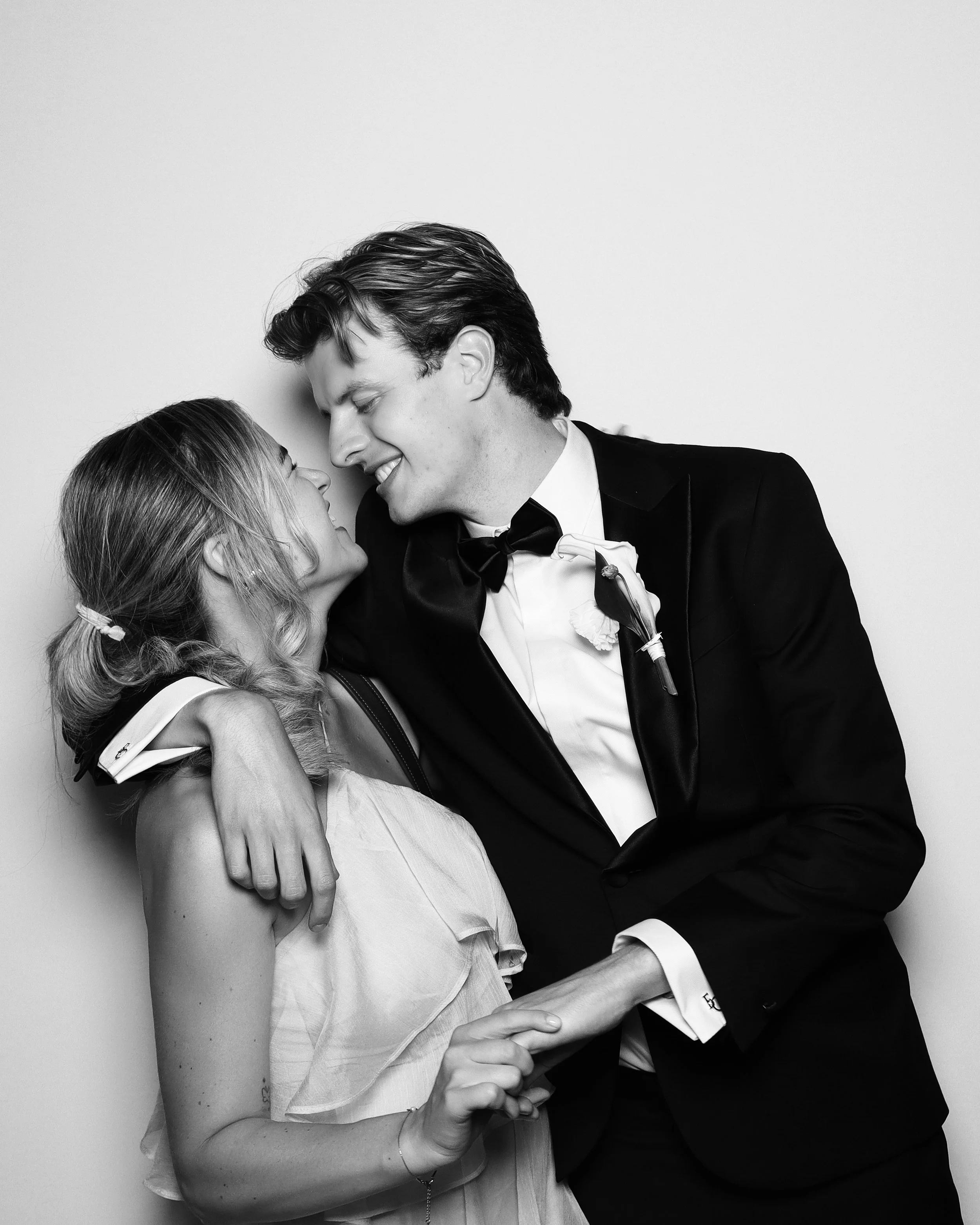 A black and white photo of a man and woman smiling and holding hands in a close embrace, looking into each other's eyes, dressed formally, likely at a wedding or special event.