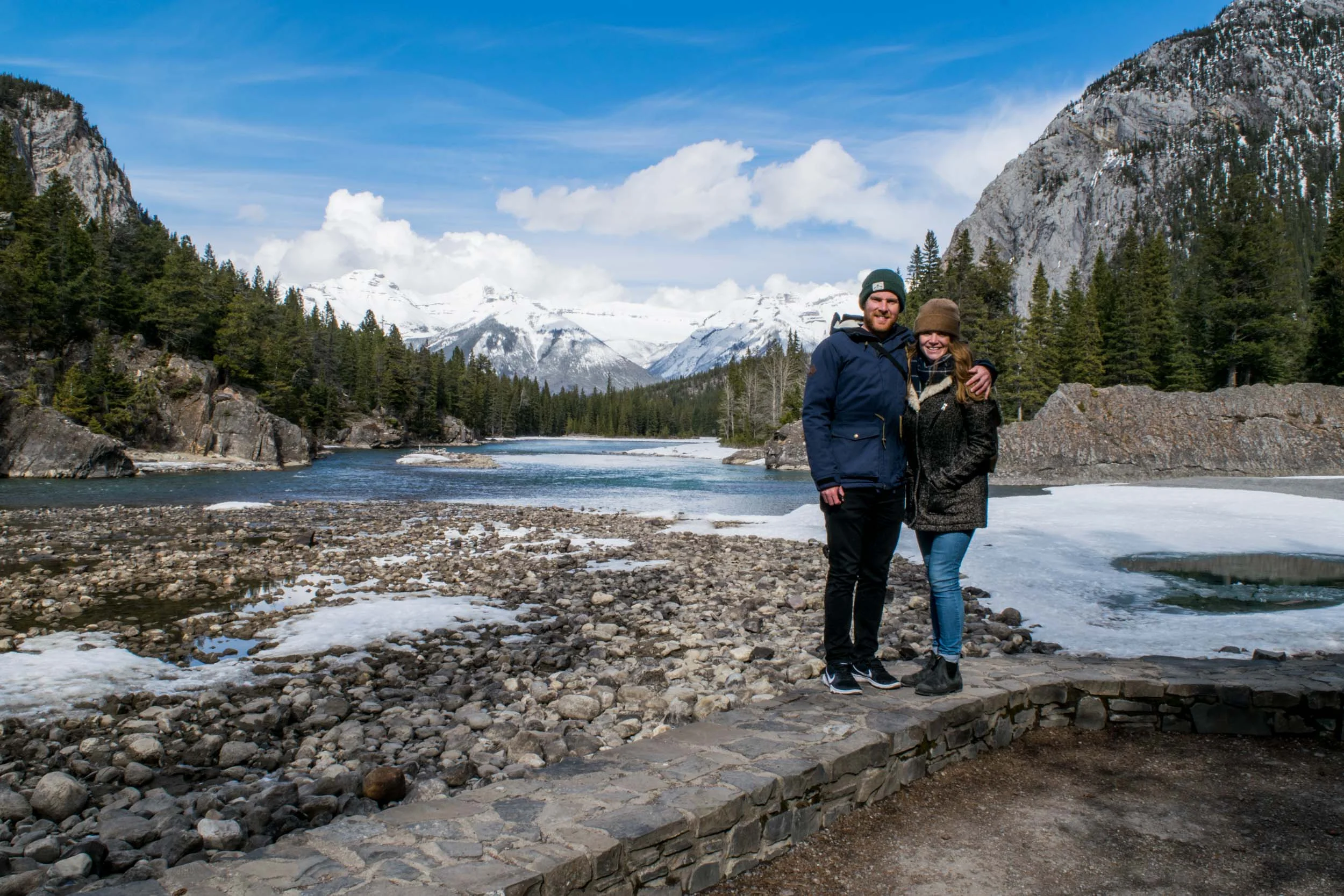 Couple stood beside the Bow River with Tunnel Mountain behind. Banff, Alberta, Canada.