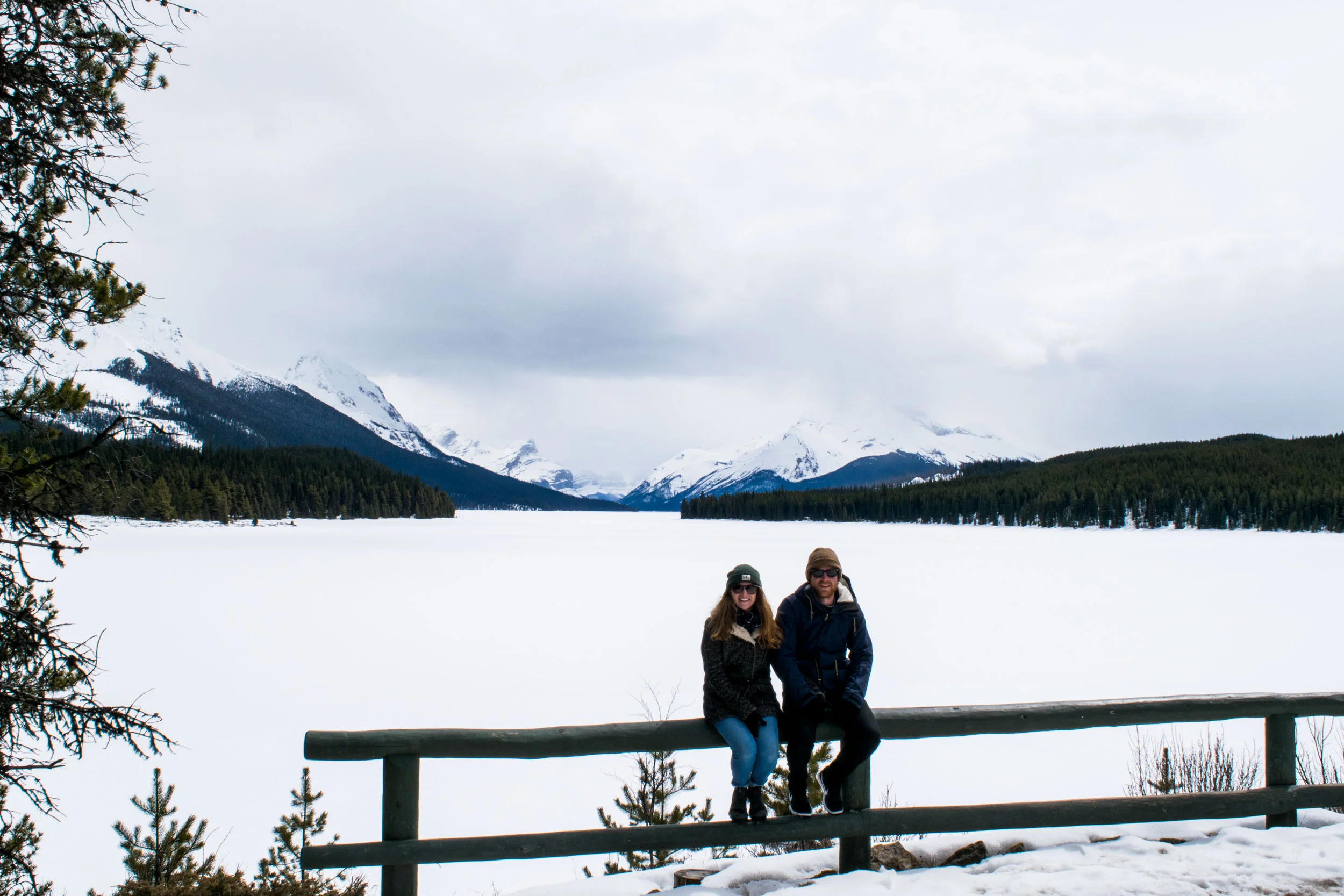 Couple sitting on a fence with a frozen and snowy Maligne Lake behind. Jasper National Park, Alberta, Canada.