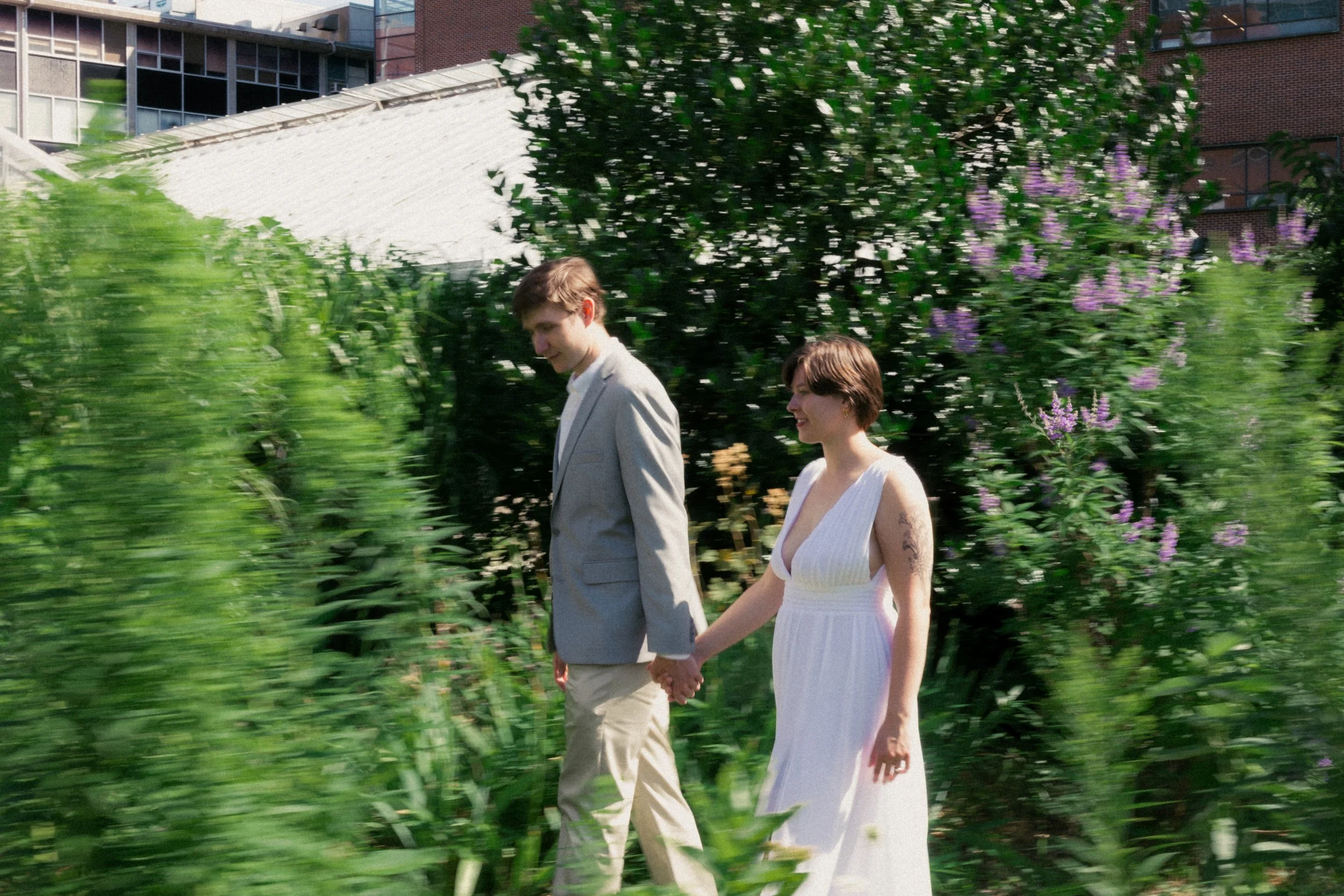 A couple holding hands walks through a lush garden with tall green bushes and purple flowers. hayden budd photo + film new england film photographer
