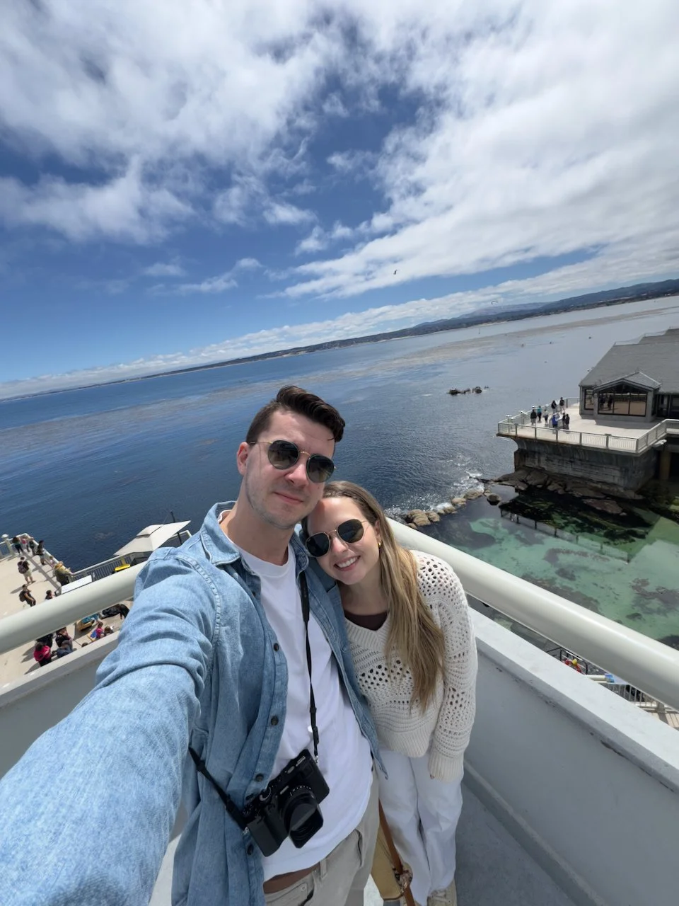 hayden budd photo + film new england film photographer Two people taking a selfie on a balcony overlooking the water, with a cloudy sky and distant shoreline in the background.