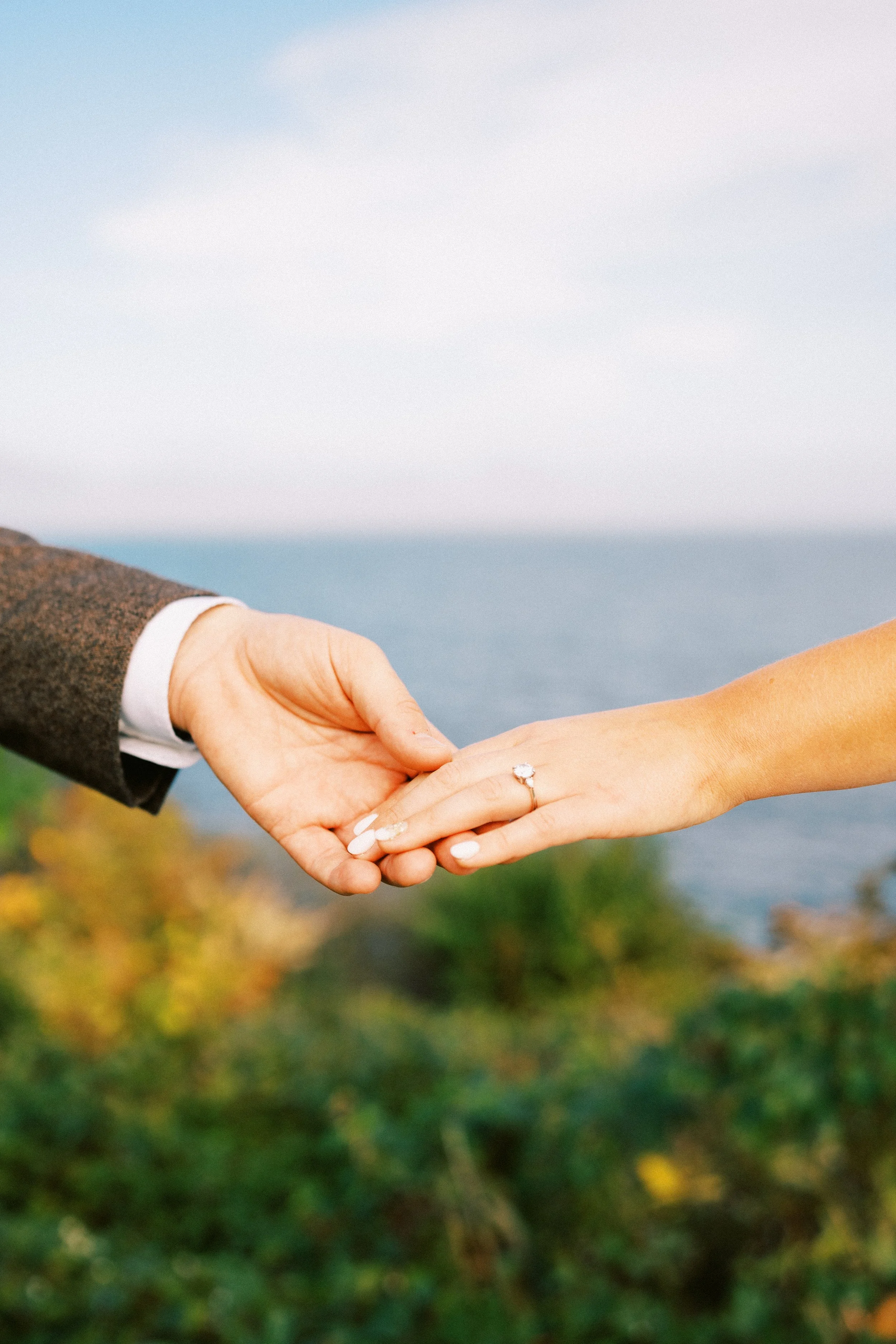 A person with a wedding ring holding hands with another person outdoors, with a body of water and greenery in the background. hayden budd photo + film new england film photographer