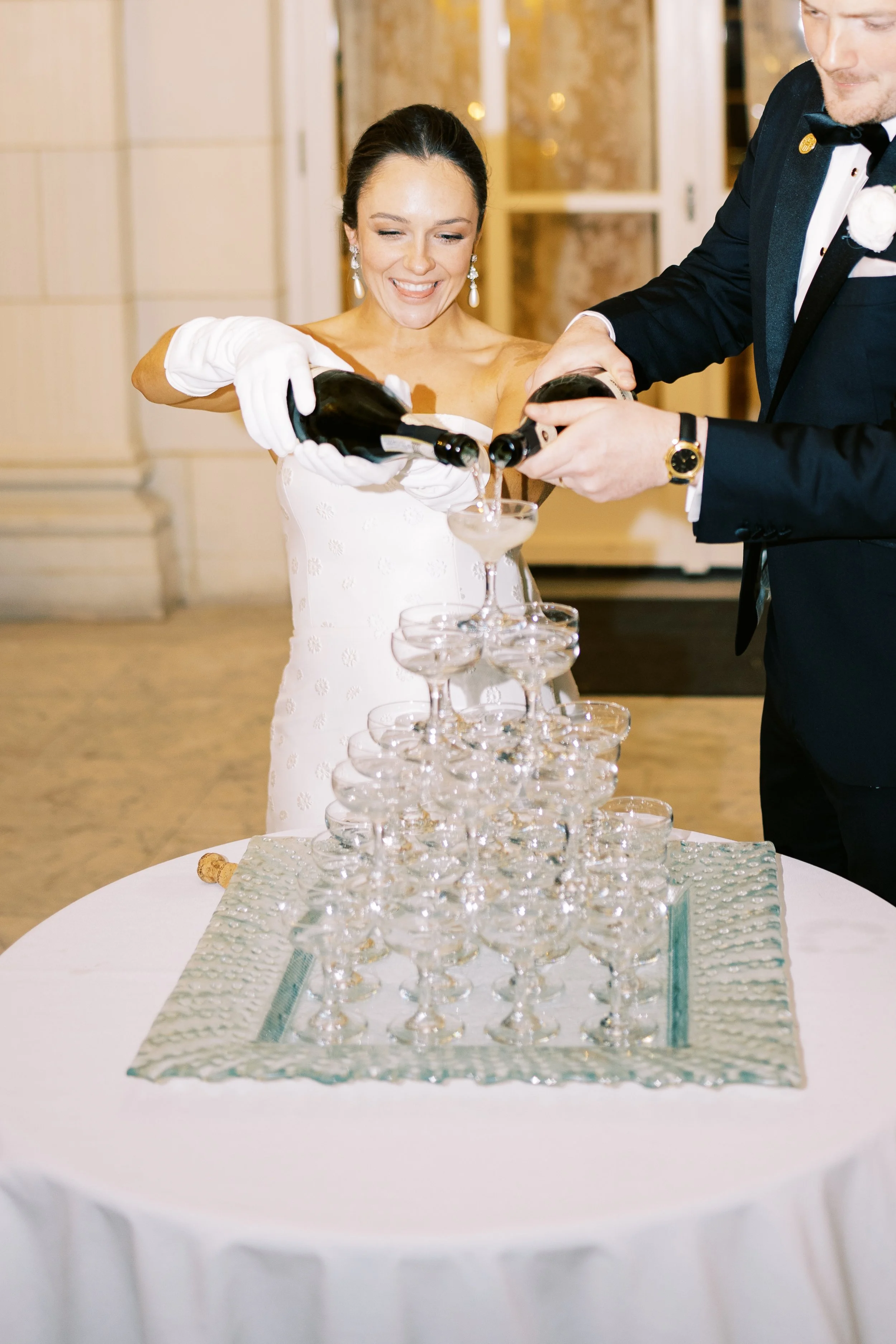 A bride in a white dress and a groom in a tuxedo pour champagne into a tower of glasses at a wedding reception. hayden budd photo + film new england film photographer