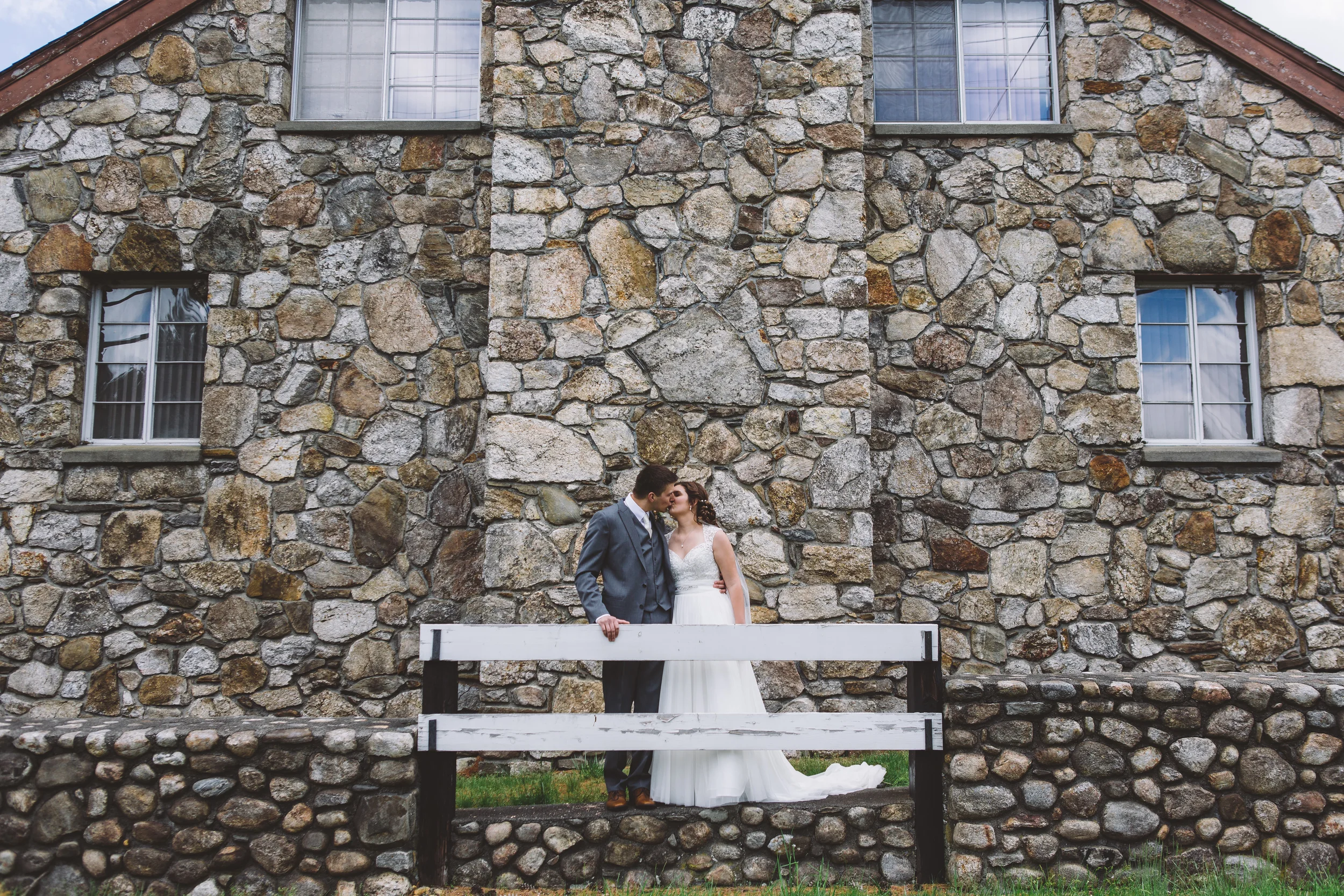 A couple in wedding attire sharing a kiss in front of a rustic stone building with four windows.