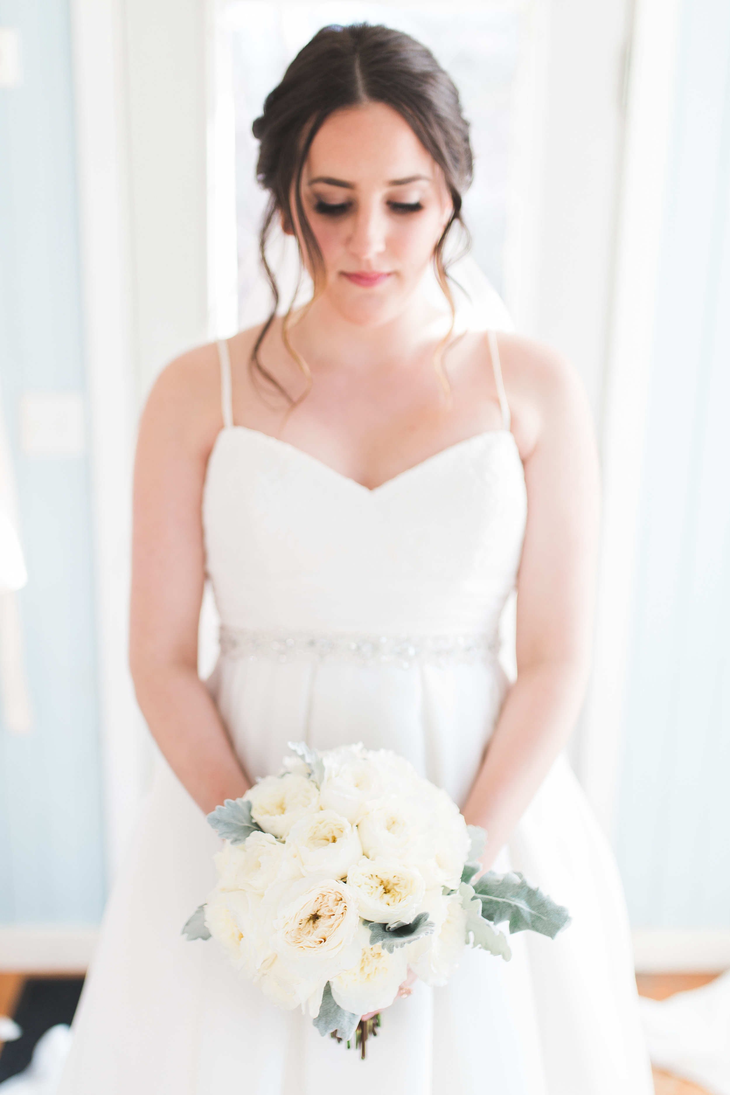 A bride in a white wedding dress holding a bouquet of white flowers, standing indoors near a window. hayden budd photo + film new england film photographer