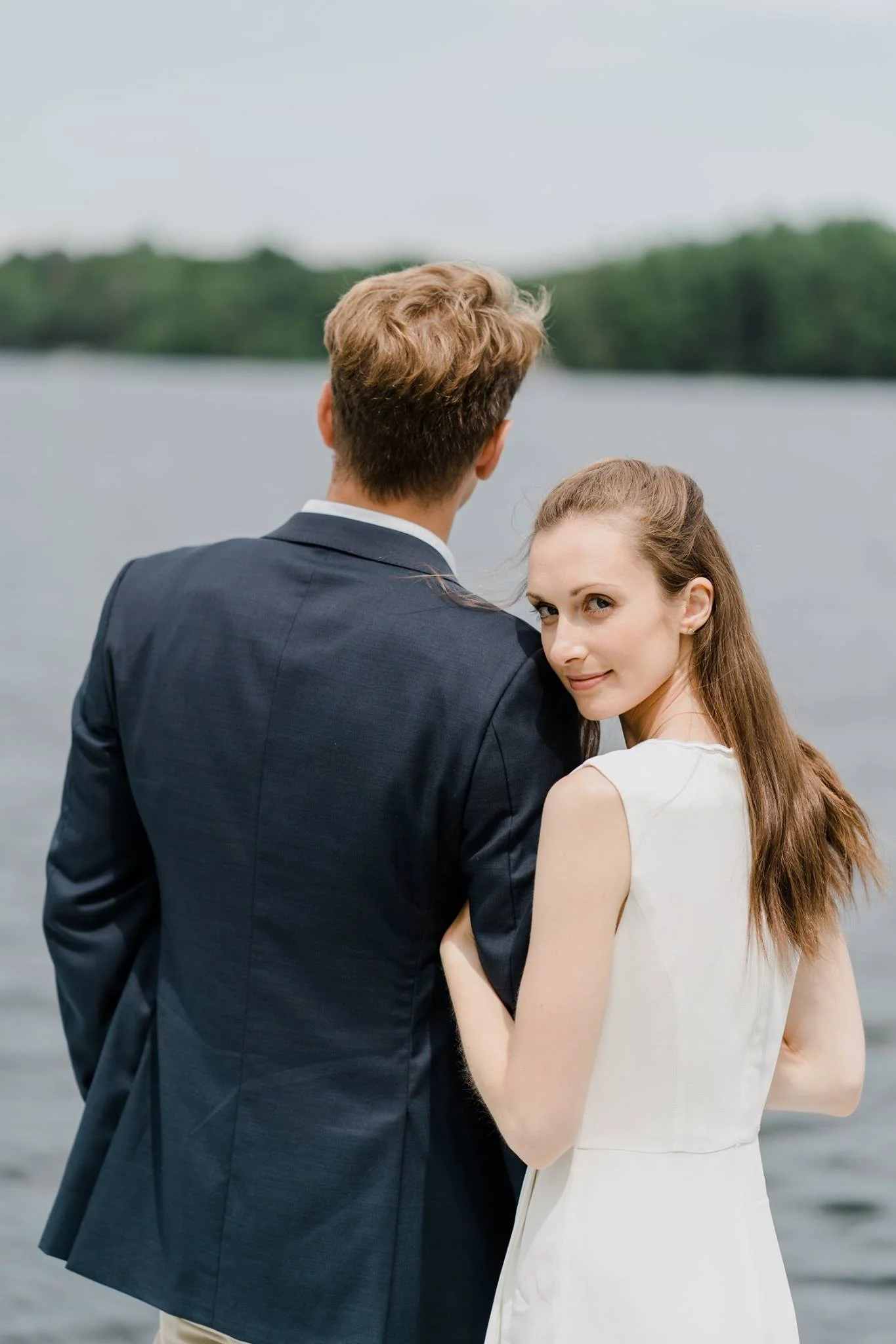 A woman with long brown hair in a white dress standing close to a man in a dark suit by a lake, with the woman looking at the camera. hayden budd photo + film new england film photographer