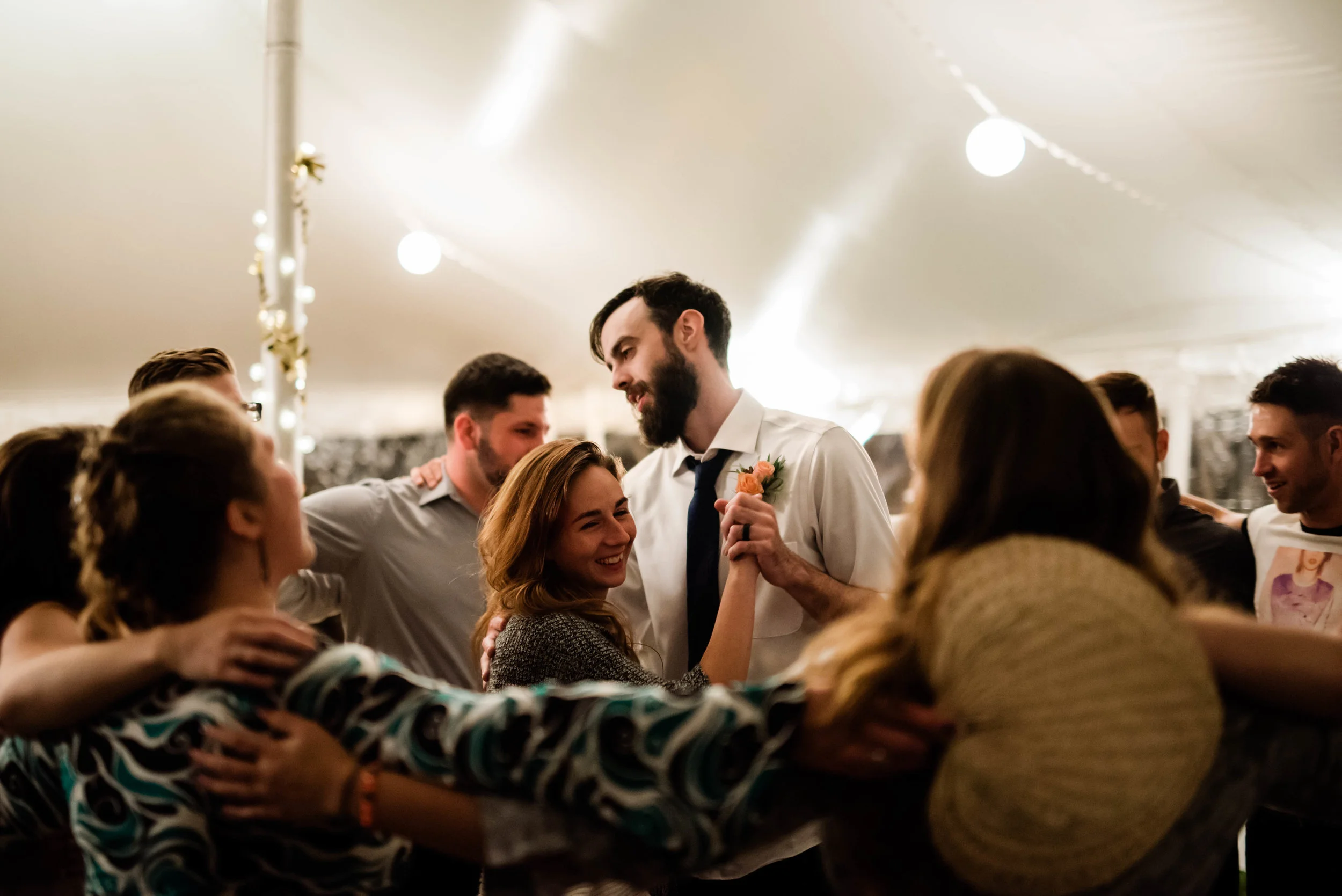 People dancing and celebrating at a party or wedding reception, with a group of friends in a circle under a tent with string lights. hayden budd photo + film new england film photographer