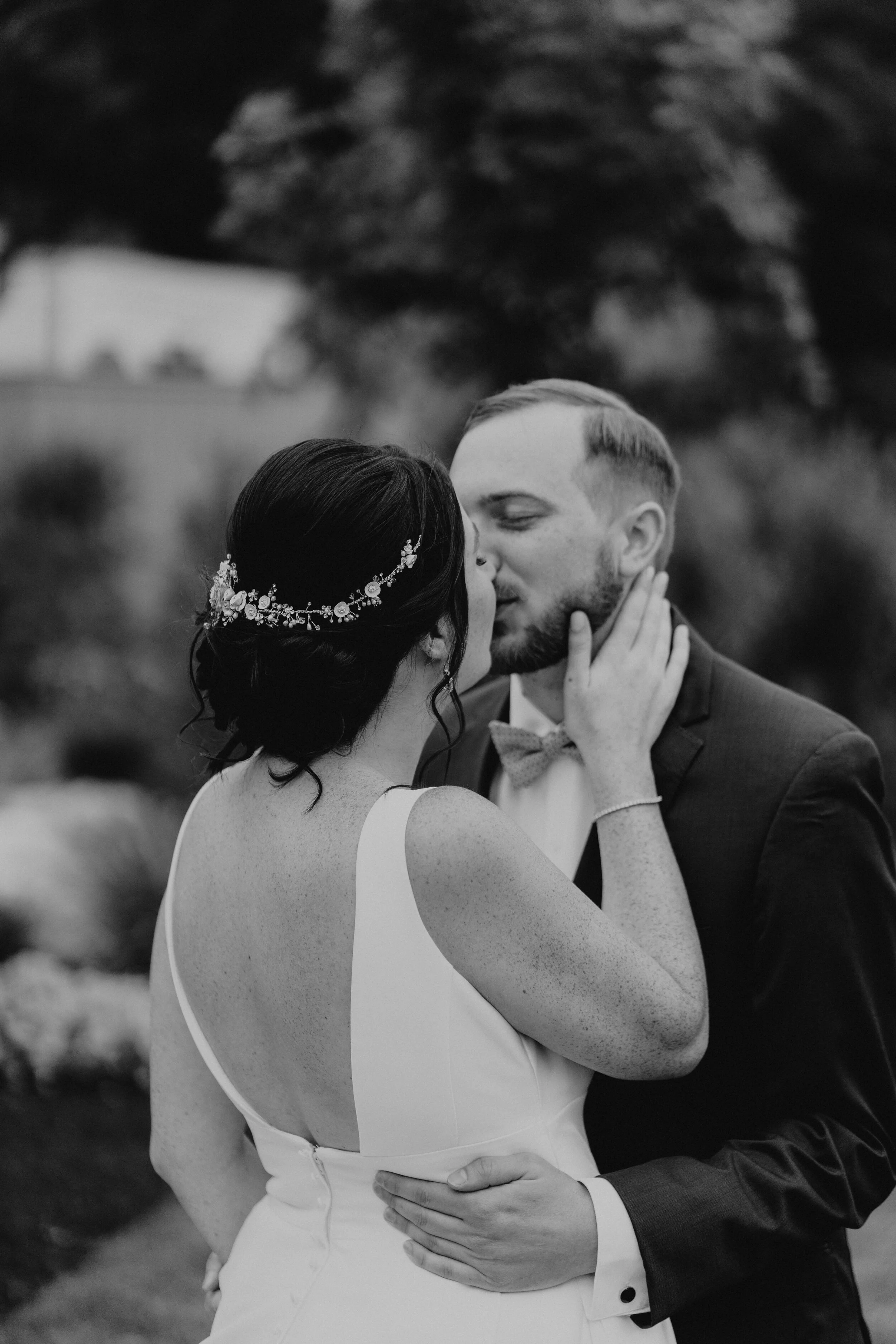 A bride and groom kissing outdoors, with the bride wearing a white dress and decorative headband, and the groom in a suit and bow tie, during a wedding ceremony or photoshoot. hayden budd photo + film new england film photographer