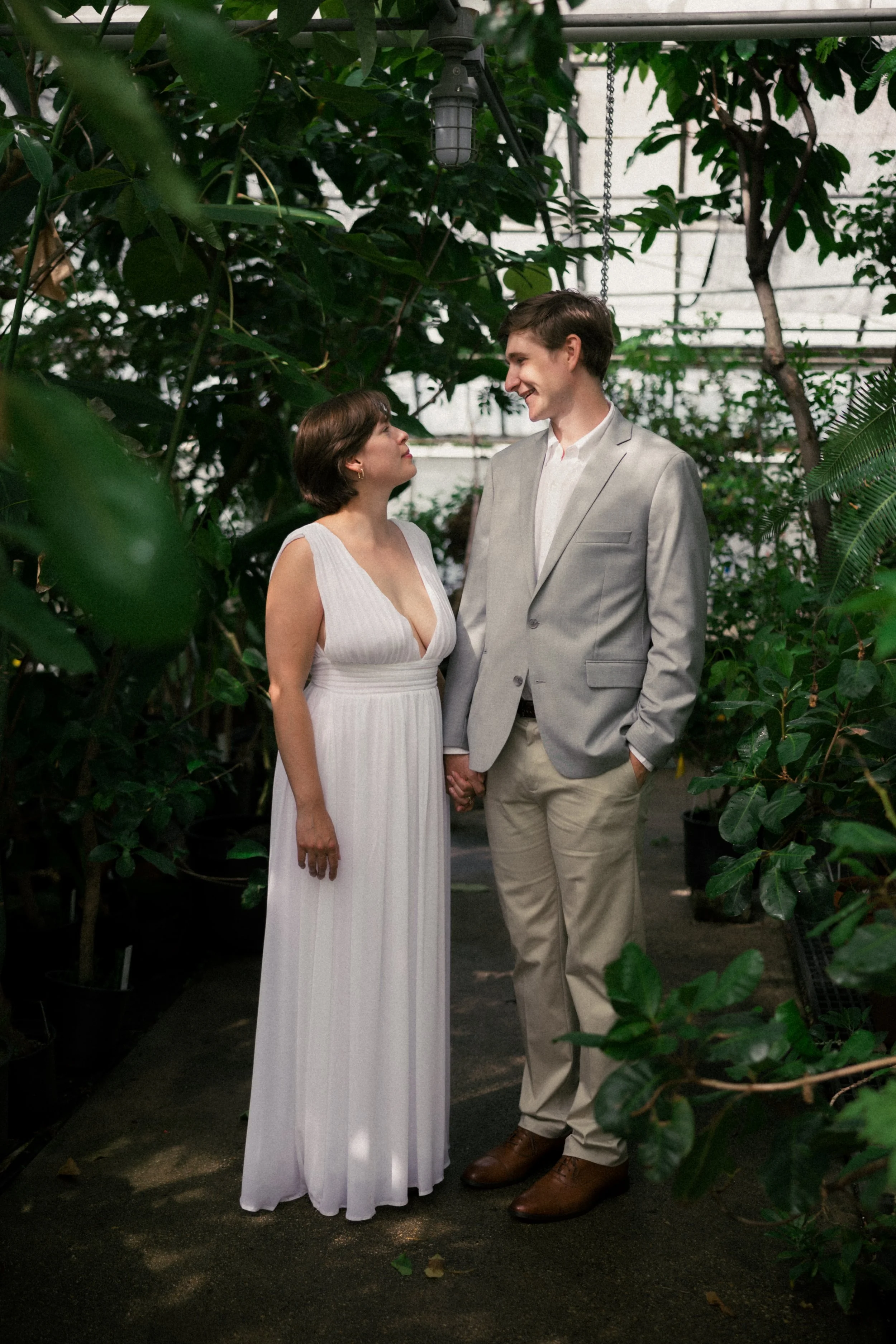 A woman in a white sleeveless dress and a man in a gray blazer and beige pants holding hands and looking at each other in a greenhouse with green plants surrounding them. hayden budd photo + film new england film photographer