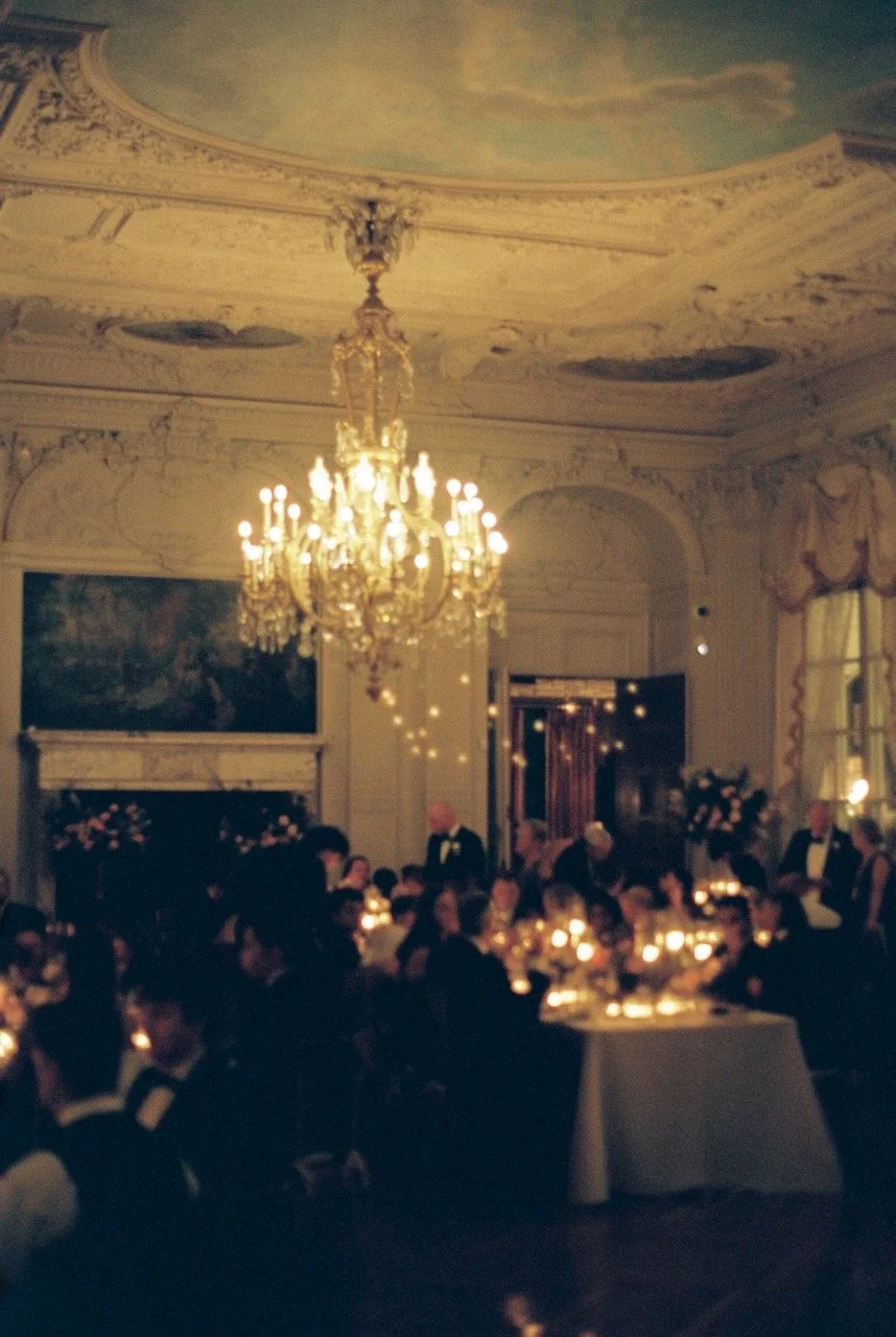 Elegant dining room with a large chandelier, decorated ceiling, and many people dressed in formal attire attending a banquet or reception with candlelit tables. hayden budd photo + film new england film photographer