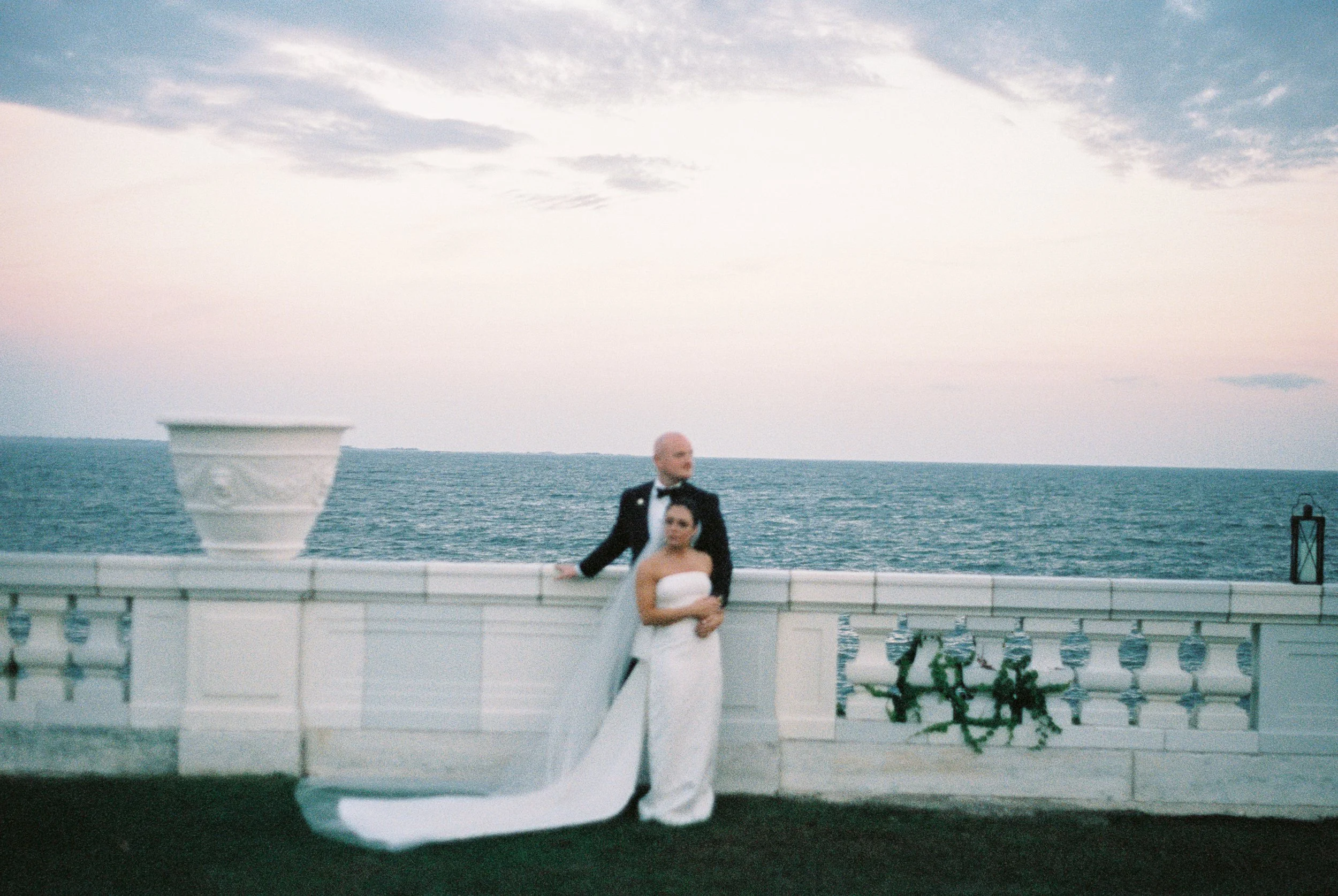 A bride and groom in wedding attire standing on a white balcony by the ocean at sunset, with a pastel sky and a decorative urn on the side. hayden budd photo + film new england film photographer
