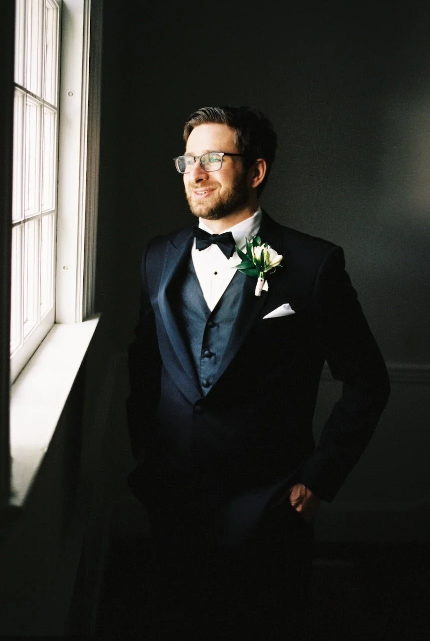 A man in a tuxedo with a boutonniere, standing by a window and looking outside with a smile. hayden budd photo + film, hayden budd photo + film new england film photographer