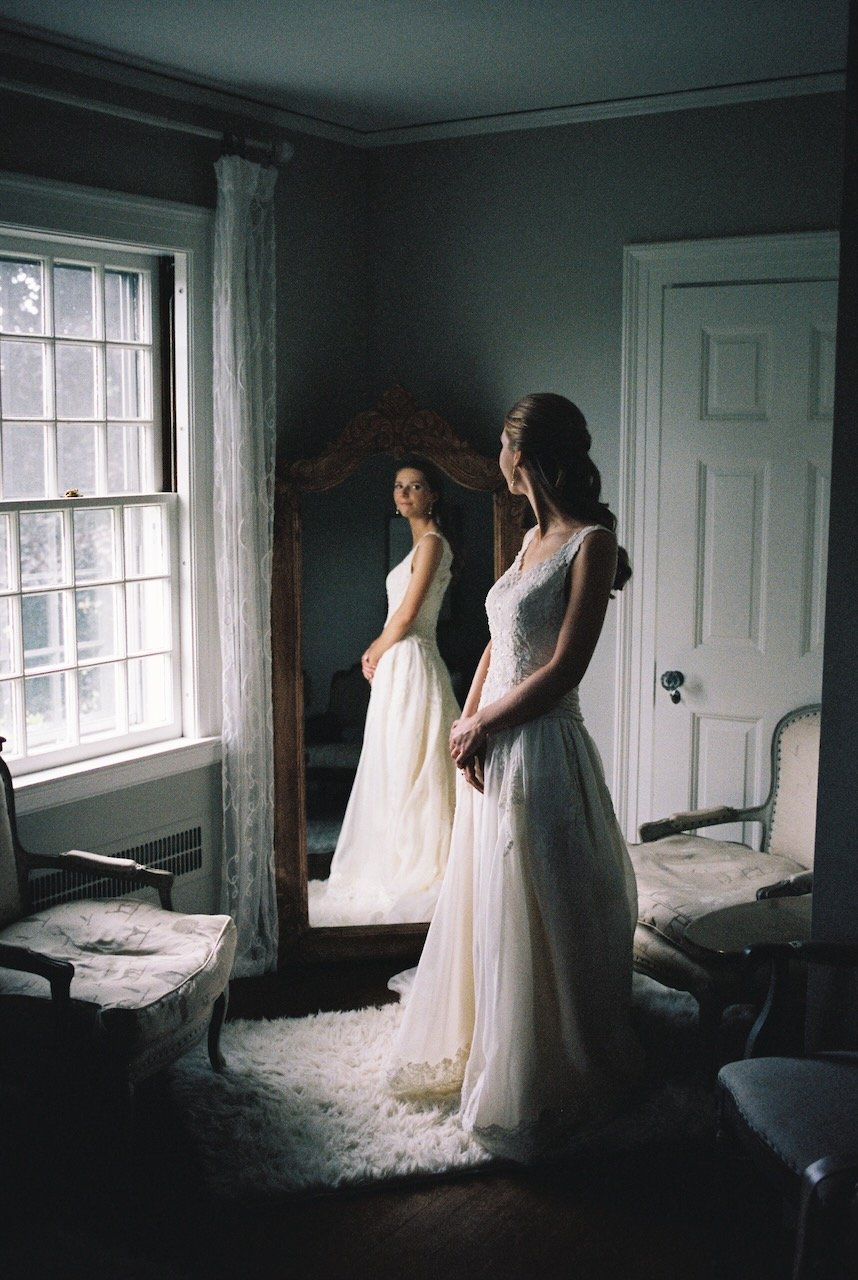 A woman in a white wedding dress looking at her reflection in an ornate full-length mirror in a cozy, softly lit room with vintage furniture and a window with white curtains. hayden budd photo + film new england film photographer