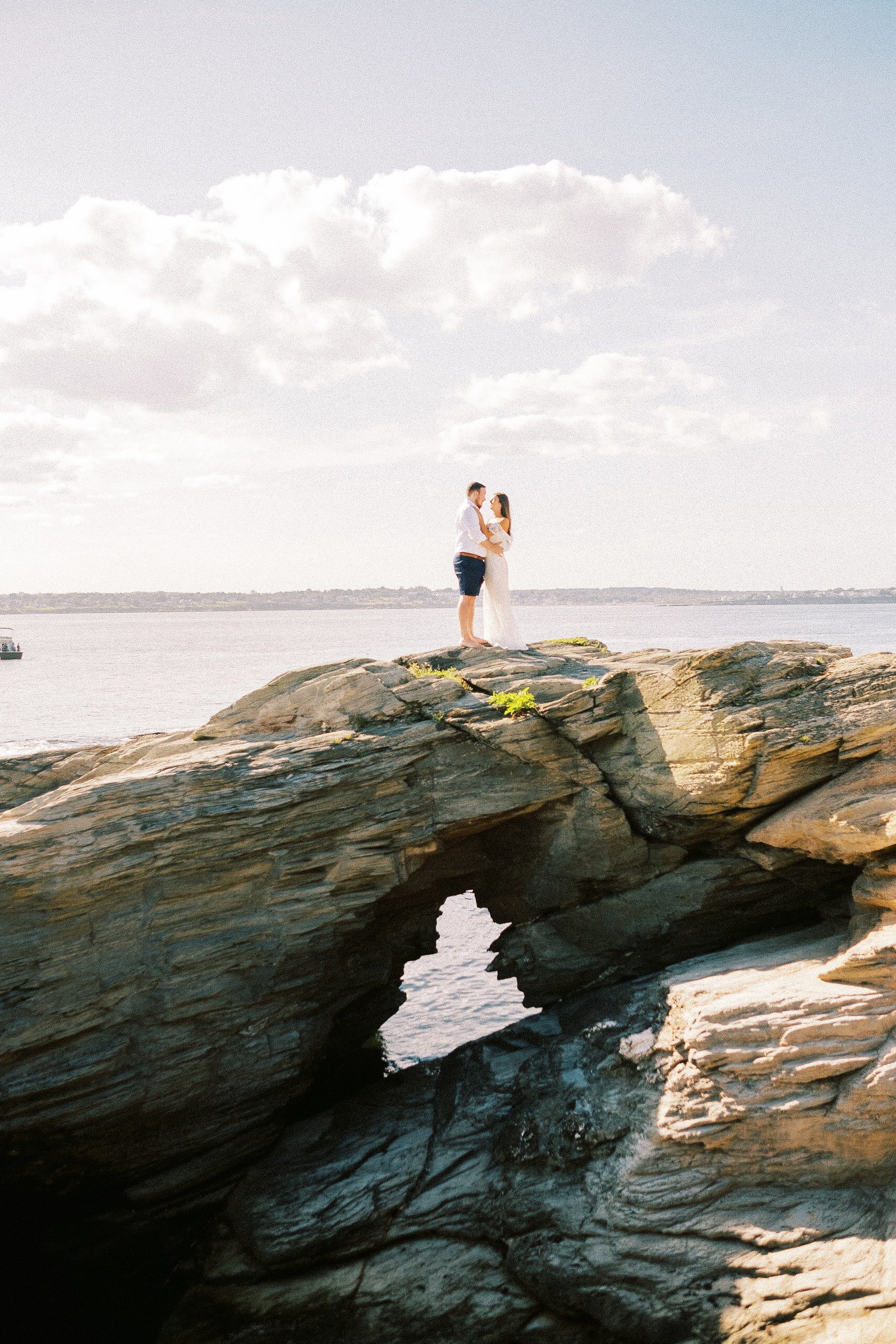 A couple stands on a rock formation near water, holding each other and gazing into each other's eyes, with a cloudy sky overhead. hayden budd photo + film new england film photographer