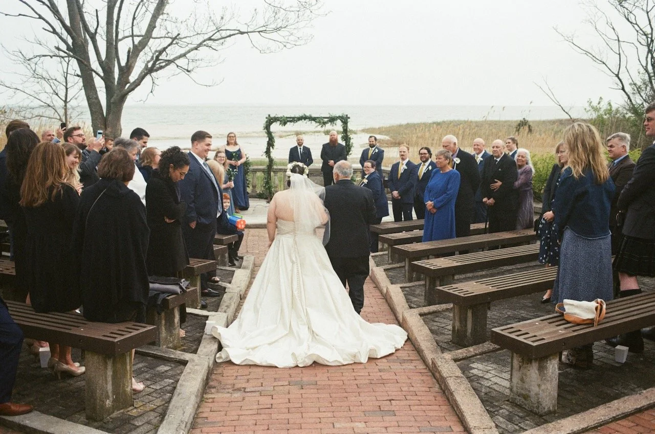 Bride walking down the aisle towards the officiant at an outdoor wedding ceremony by the water, with guests standing on either side. hayden budd photo + film new england film photographer