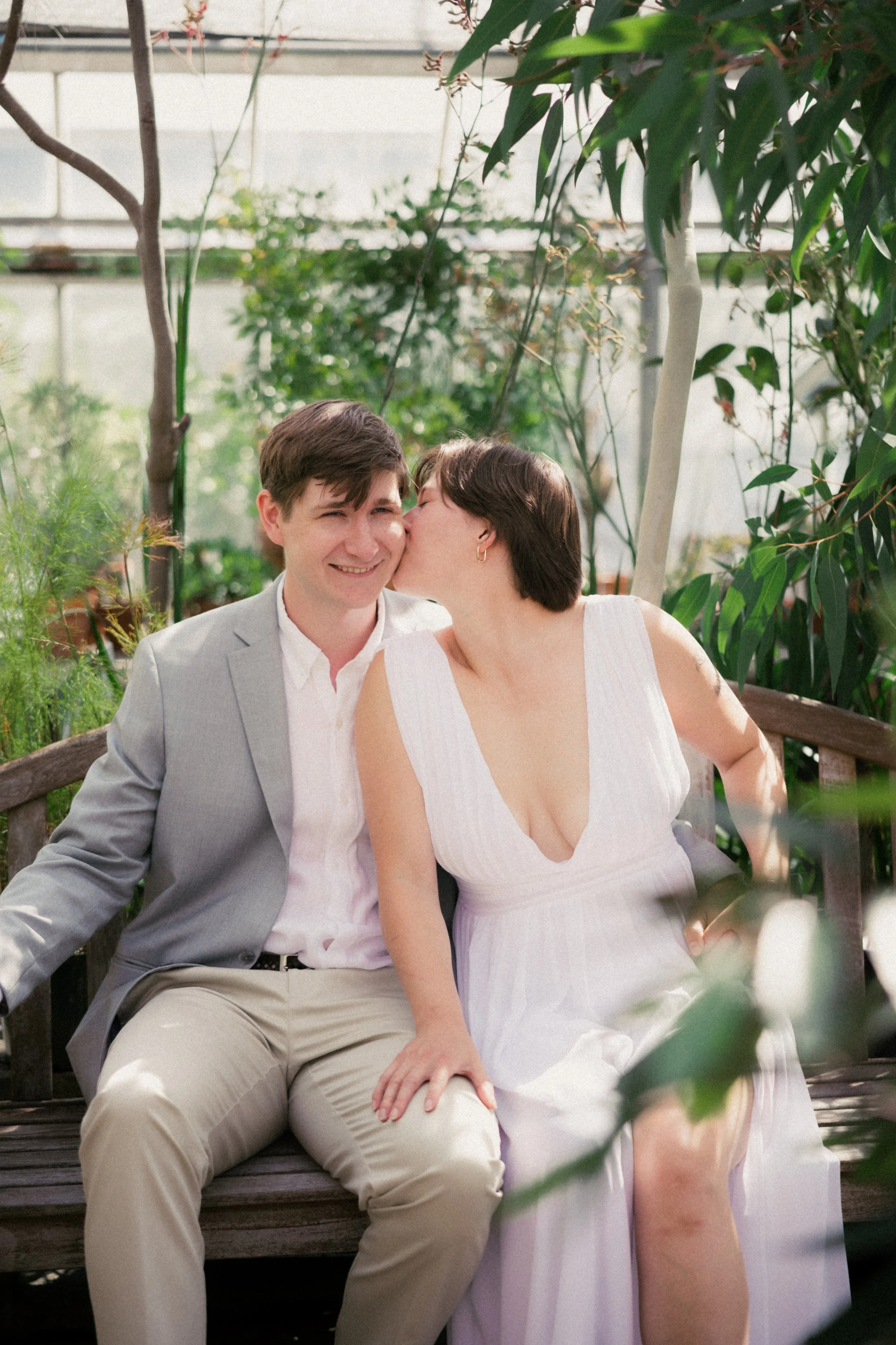A woman kisses a man on the cheek while sitting on a bench in a greenhouse filled with lush green plants. hayden budd photo + film new england film photographer