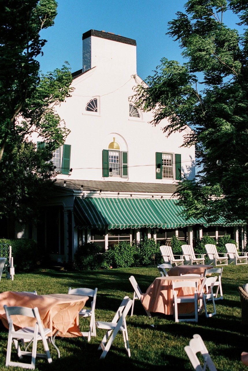 A white multi-story building with green window shutters, surrounded by trees and outdoor seating with tables and chairs. hayden budd photo + film new england film photographer