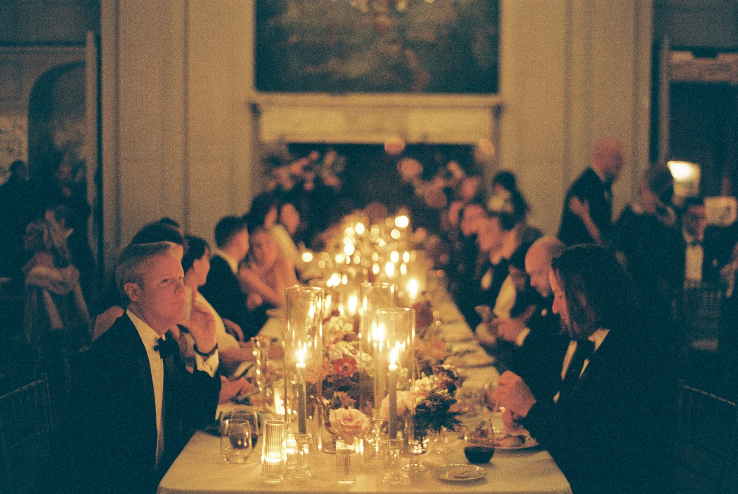 A formal dinner event with a long table decorated with candles and flowers, surrounded by people in suits and dresses, in a room with warm lighting and a fireplace in the background. hayden budd photo + film new england film photographer