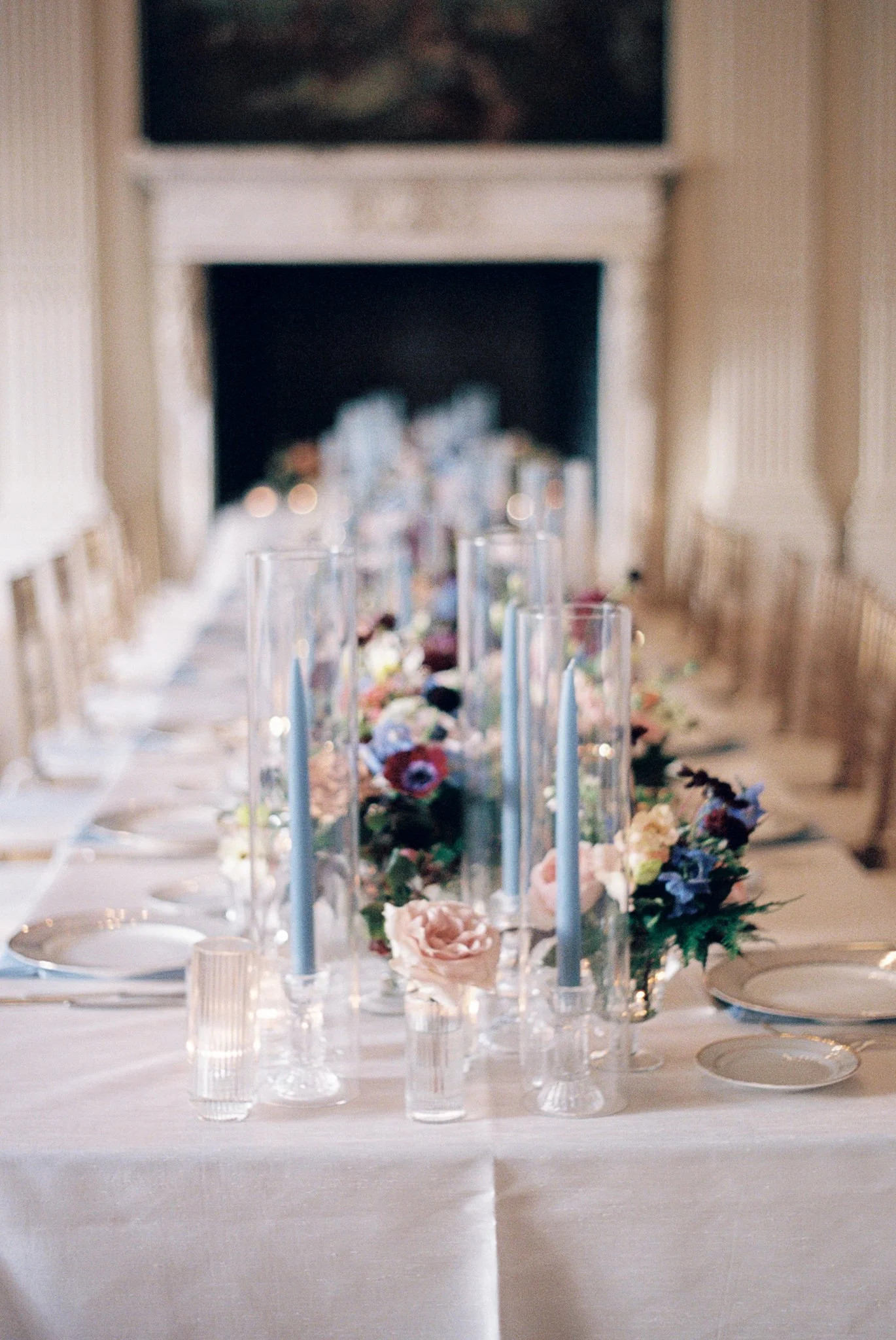 Elegant wedding reception table with floral centerpieces, tall glass candle holders, blue candles, and white tablecloth, set in a sophisticated room with a fireplace. hayden budd photo + film new england film photographer