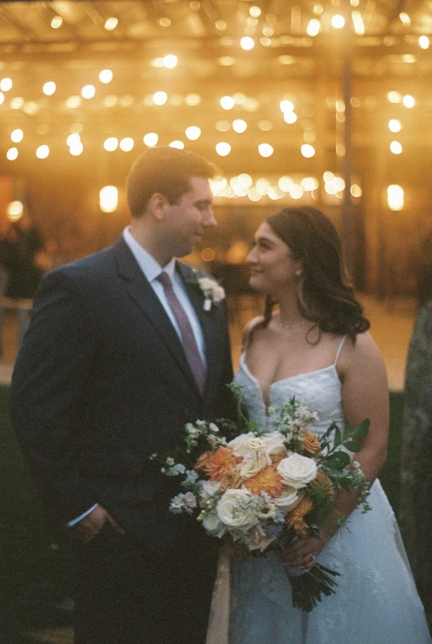 A bride and groom stand close together holding a bouquet of flowers, with warm, blurred string lights overhead, at a wedding reception. hayden budd photo + film new england film photographer